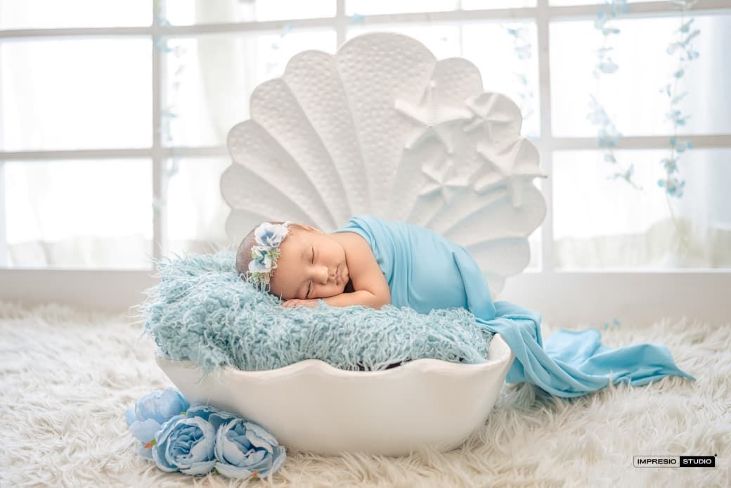 Newborn baby sleeping peacefully in a white shell bowl with blue wrap and floral headband on fluffy white rug.