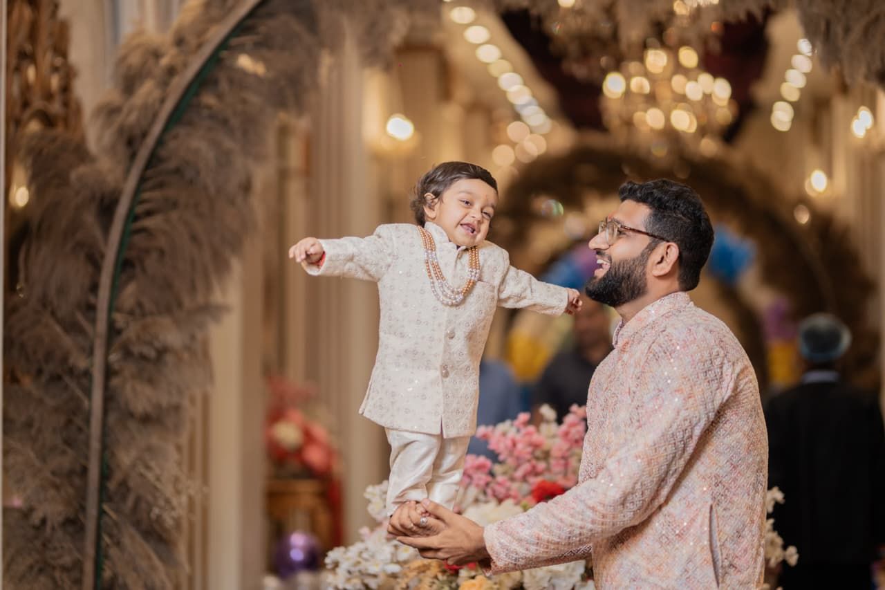 Man in pink kurta lifts laughing toddler in cream sherwani at elegant Indian wedding celebration.