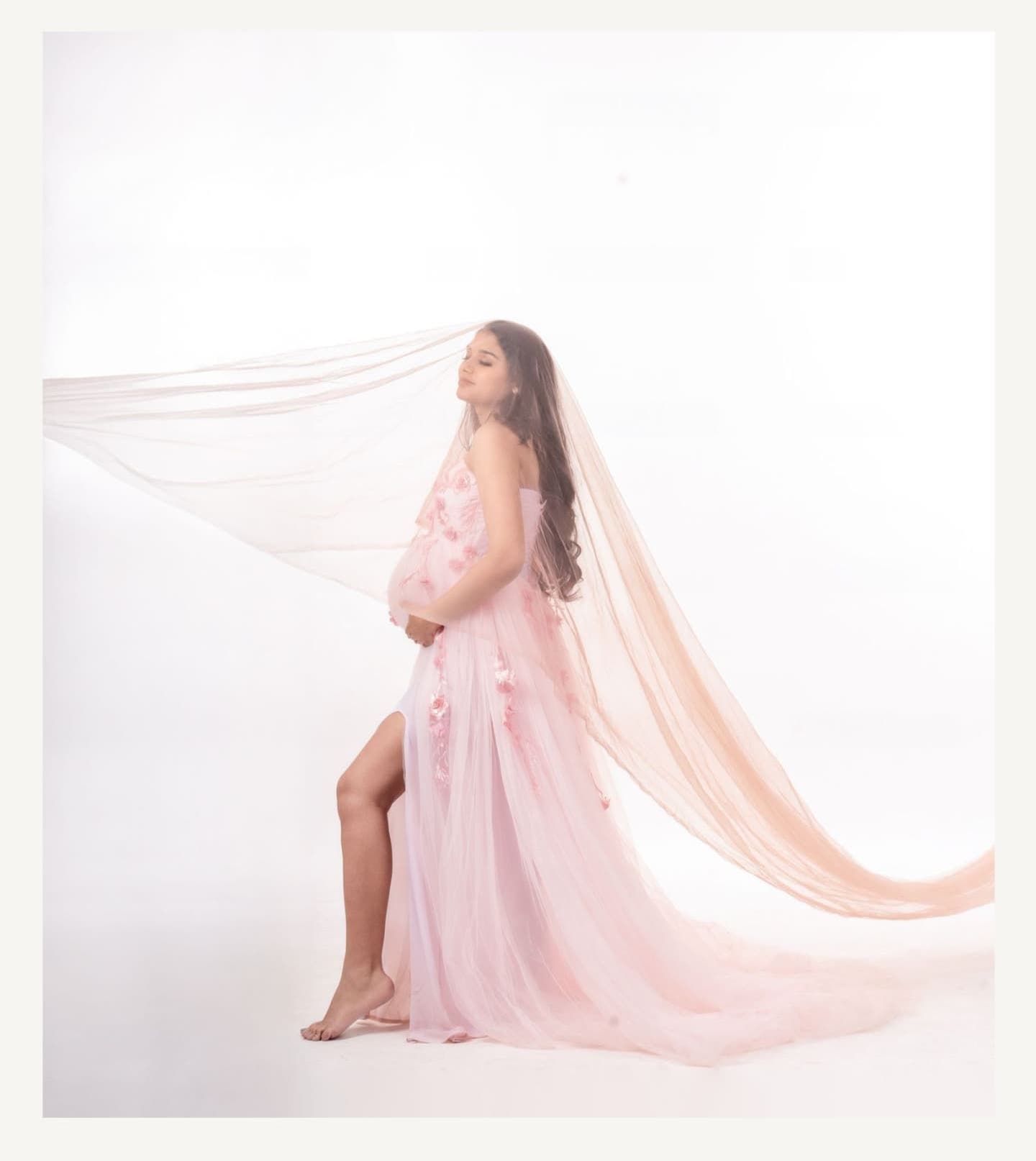 Ethereal maternity photoshoot featuring a flowing pink sheer dress and long veil against a white backdrop.