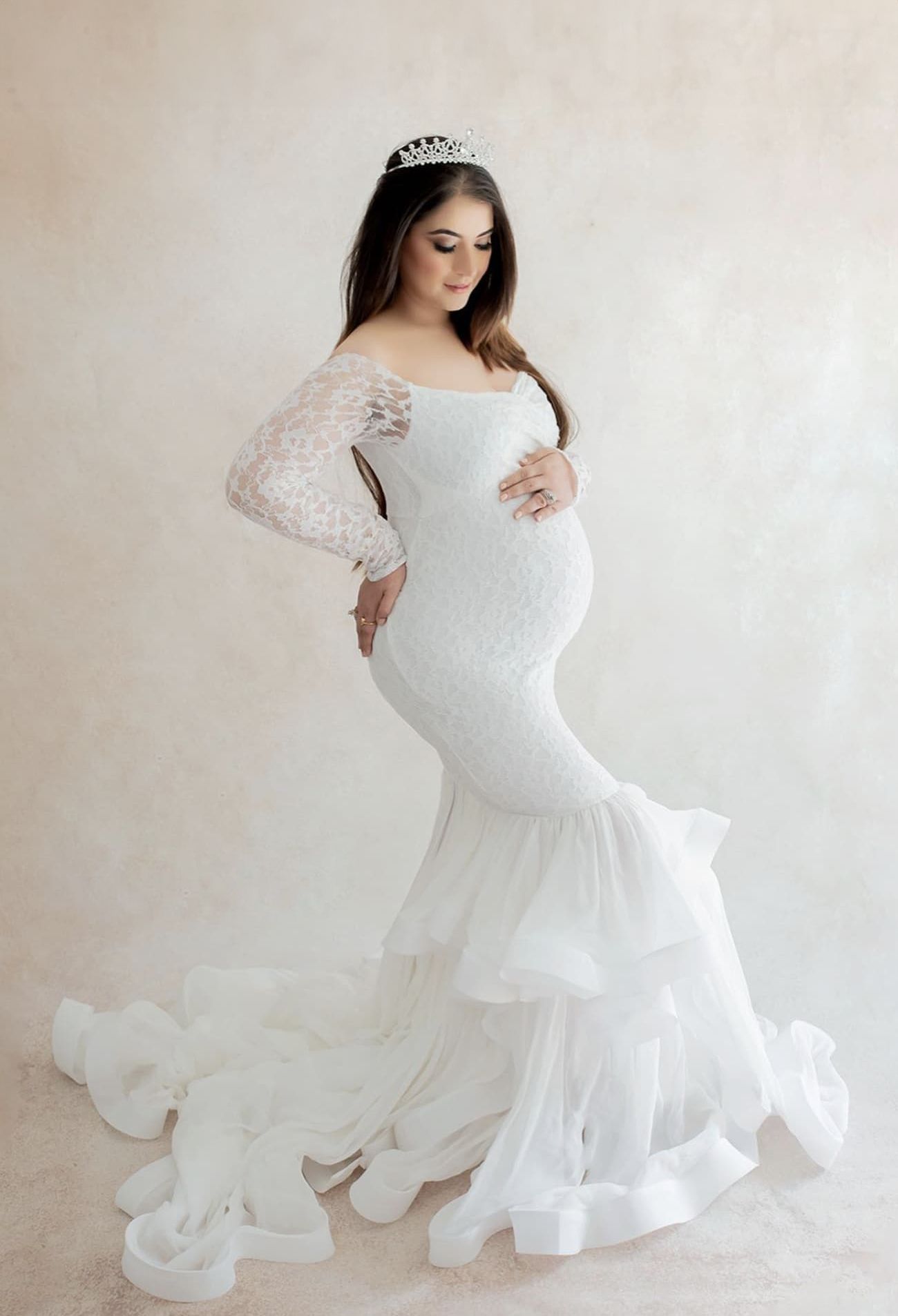 Elegant maternity portrait in white lace gown with long sleeves and mermaid silhouette against neutral backdrop.