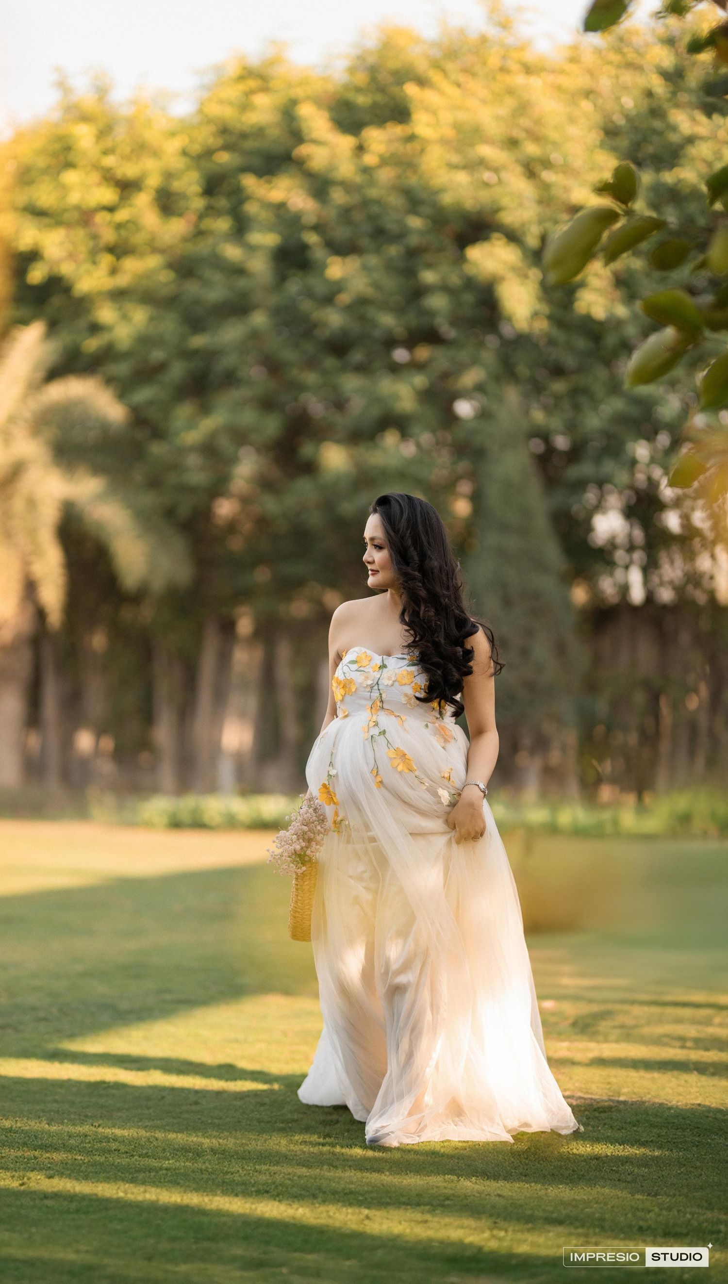 Elegant outdoor maternity photo featuring a flowing white and gold dress against a sunset-lit garden landscape.