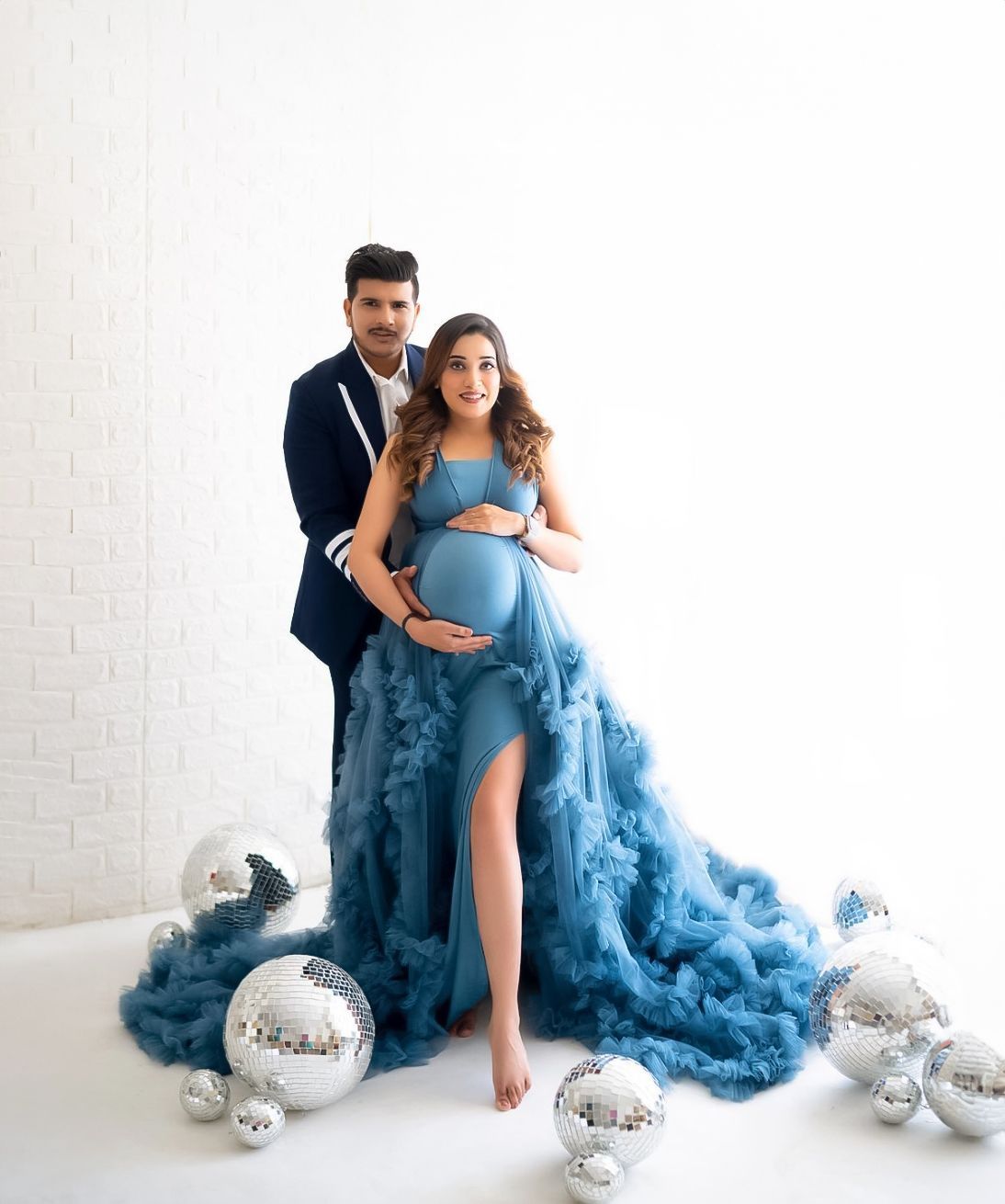 Couple's maternity portrait in blue tulle gown with silver ornaments against white wall.