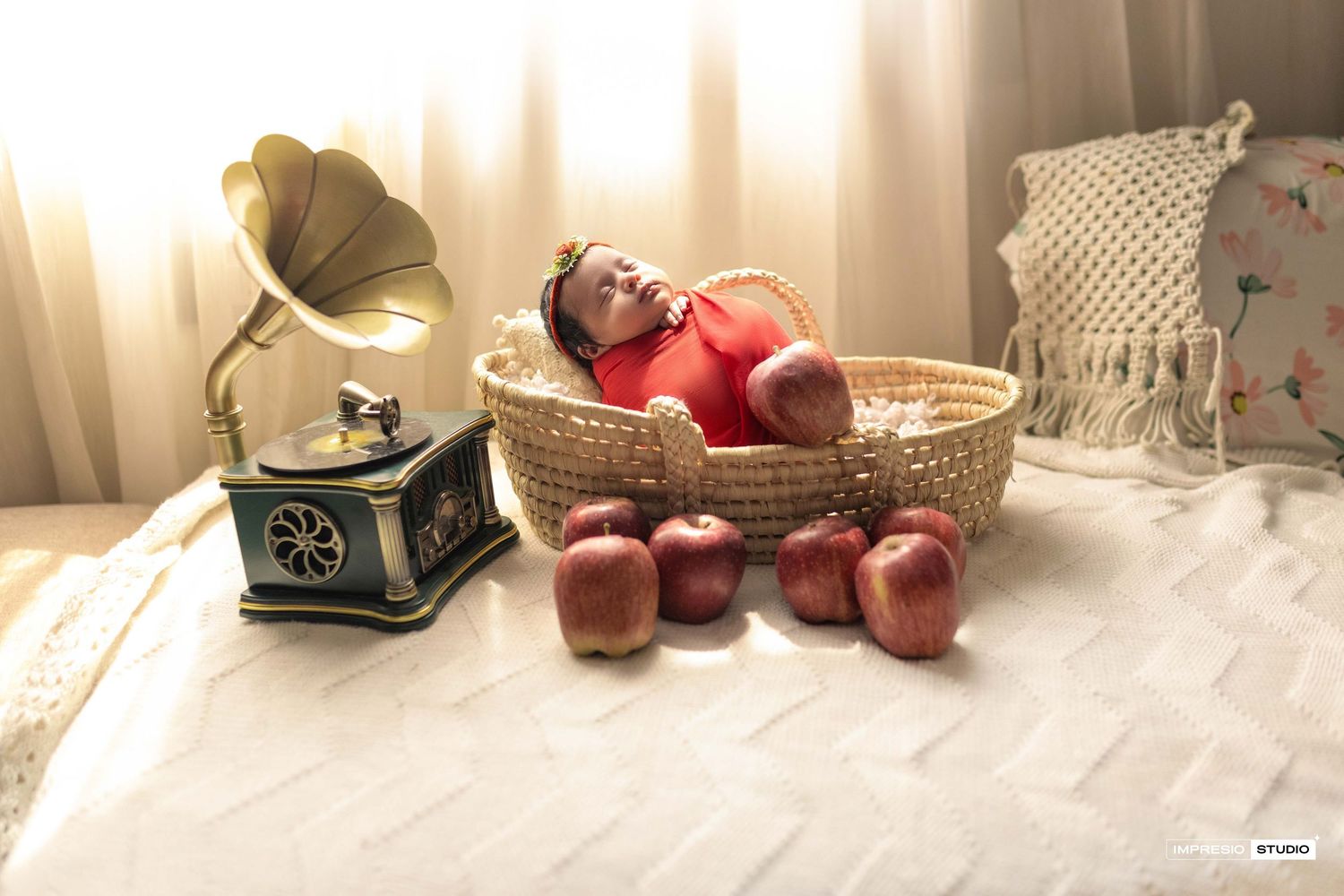 Baby sleeping in a wicker basket surrounded by red apples next to a vintage gramophone on a white bed.