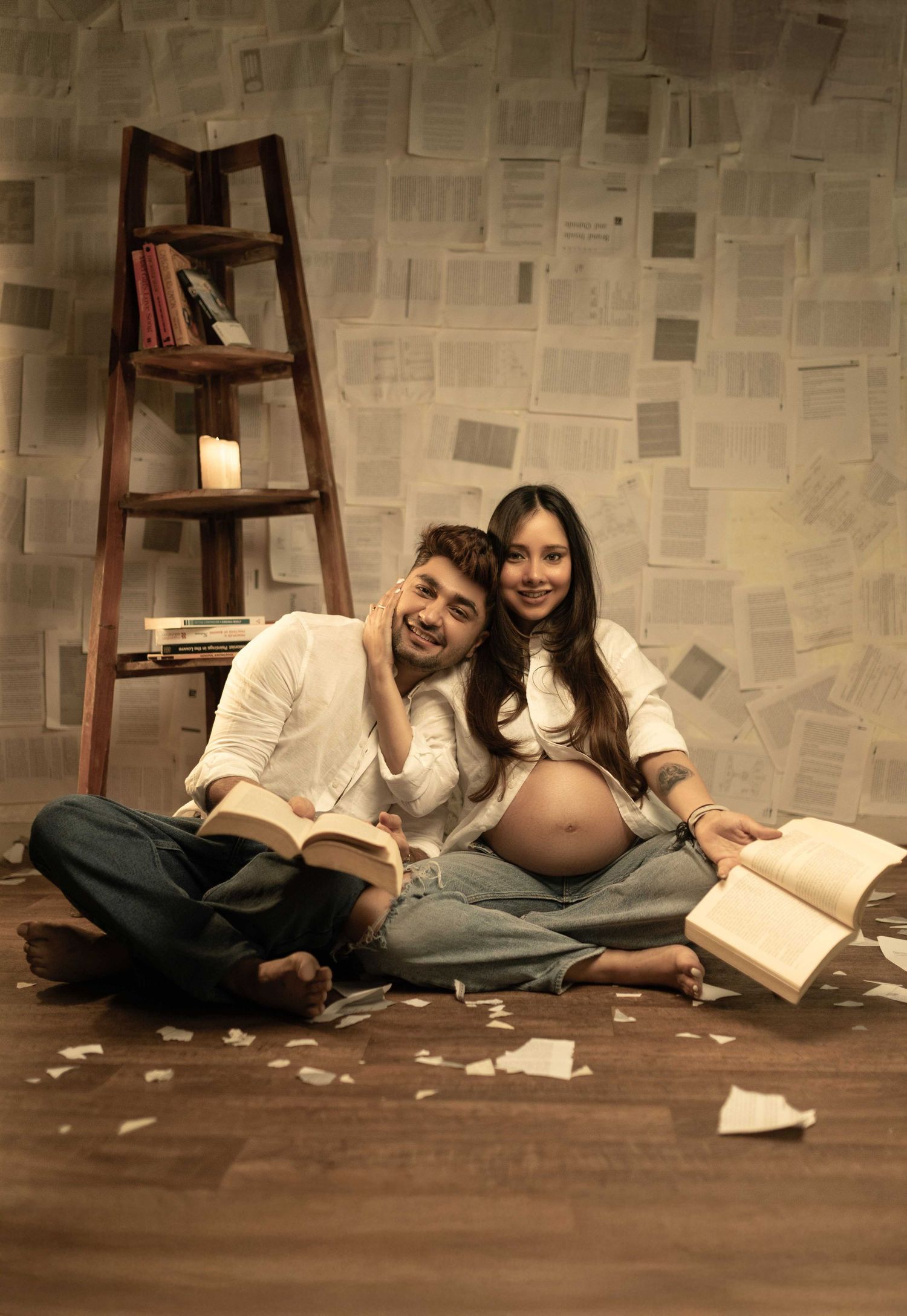 Couple sitting on floor with open books, pregnant woman showing baby bump, ladder bookshelf and pages on wall backdrop.