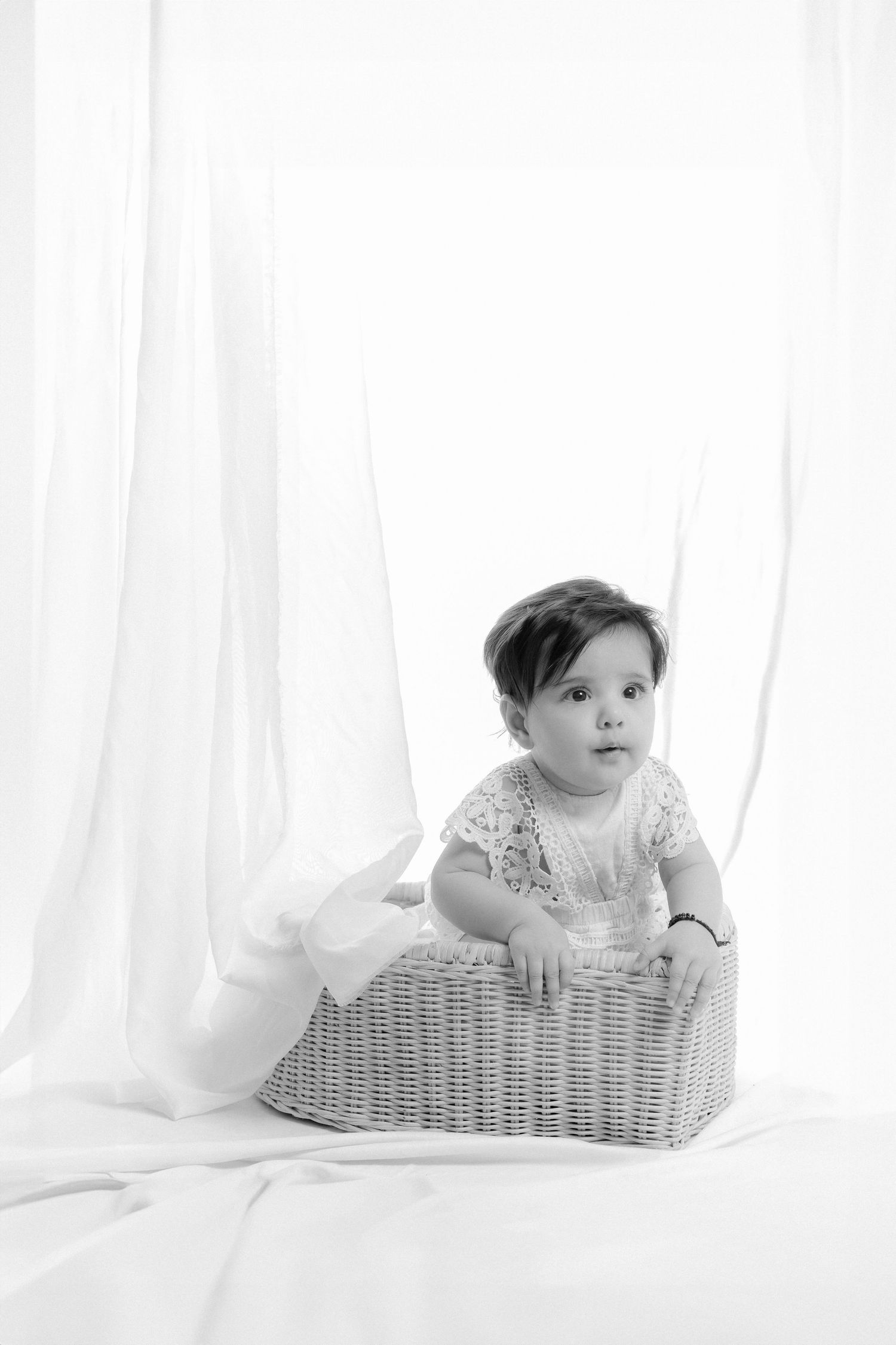 Black and white photo of a cute baby sitting in a wicker basket with white draping curtains in the background.