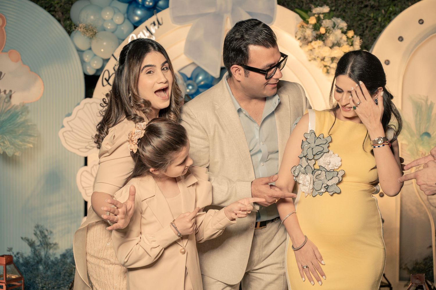 Family celebrating a baby shower with a pregnant woman in yellow dress, man in beige suit, and two girls posing together.