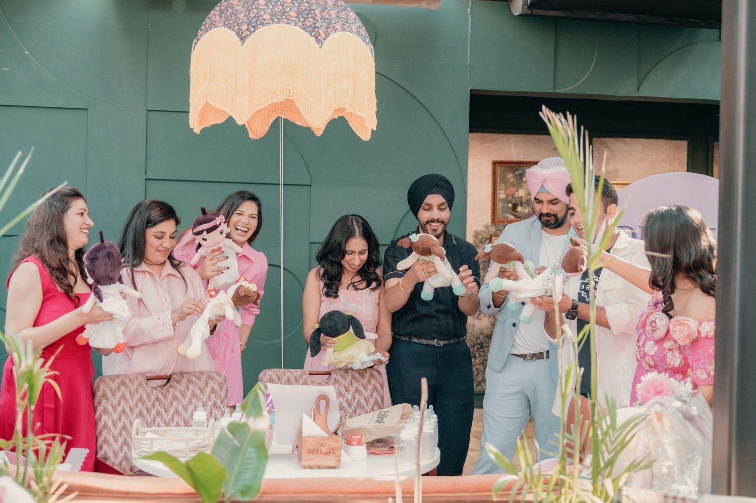 A joyful Sikh family celebration with guests holding babies outdoors, decorated with plants and a fringed umbrella.