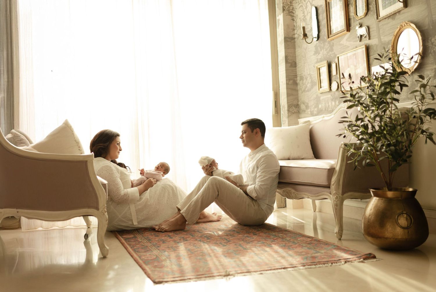 A young family sits together on a rug in a bright, elegant living room, with a mother holding a newborn baby.