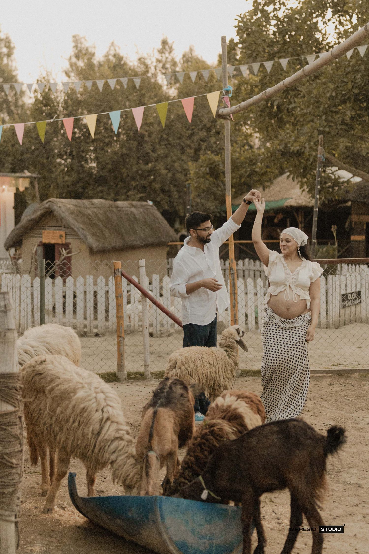 Pregnant woman and man dancing among llamas and goats at a festive outdoor farm with colorful bunting.