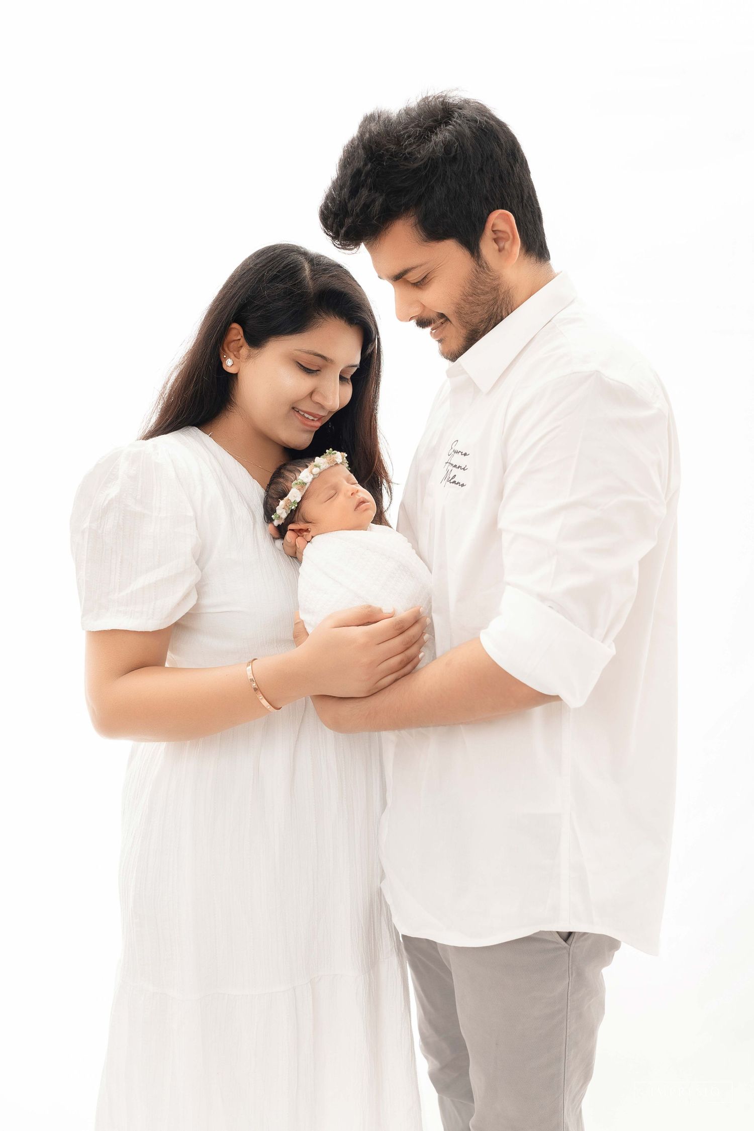 A young couple in white outfits lovingly gazes at their swaddled newborn baby against a white background.