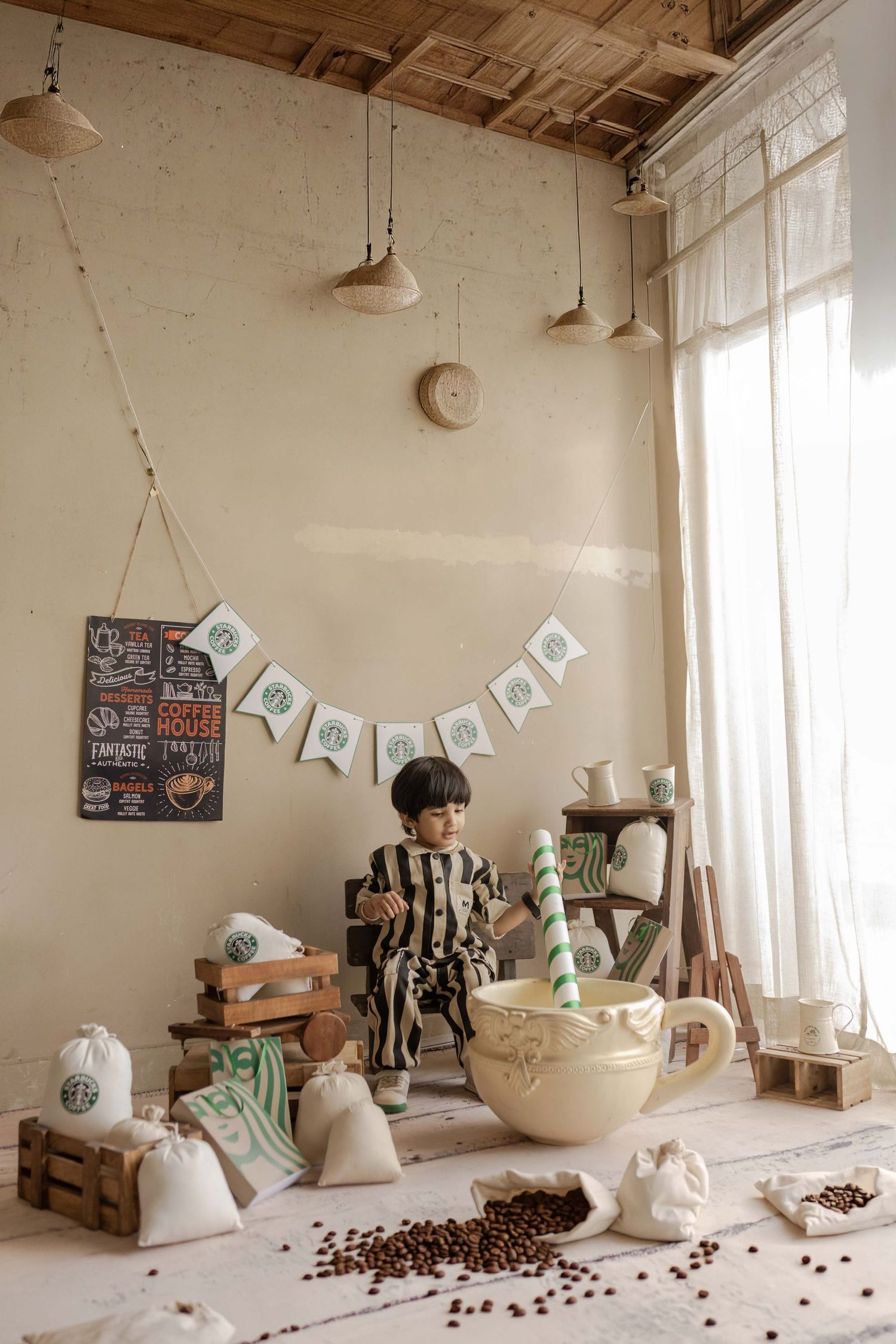 Child sits beside oversized coffee cup prop with Starbucks sacks and beans in a rustic cafe-themed photo set.