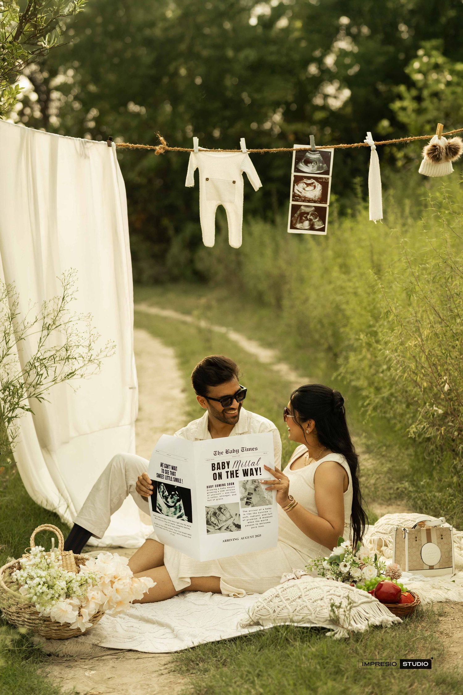 Couple sitting outdoors on a blanket for a maternity photoshoot with ultrasound photos and baby clothes on a line.