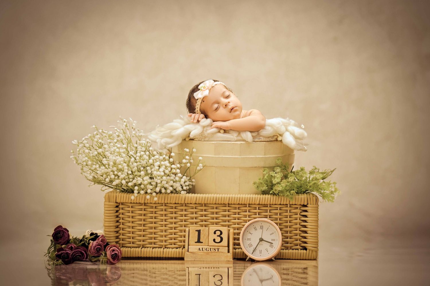 Newborn baby sleeping peacefully in a decorative basket with white flowers and a small clock showing number 13.