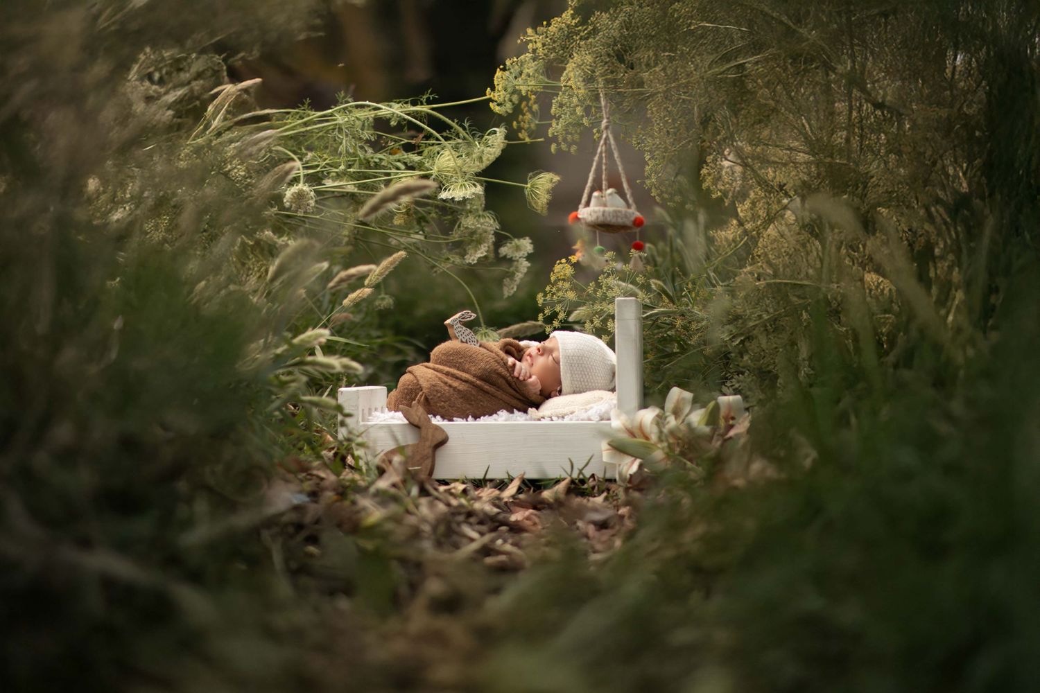 Newborn baby sleeping peacefully on a white bed surrounded by lush green foliage in an enchanted garden setting.