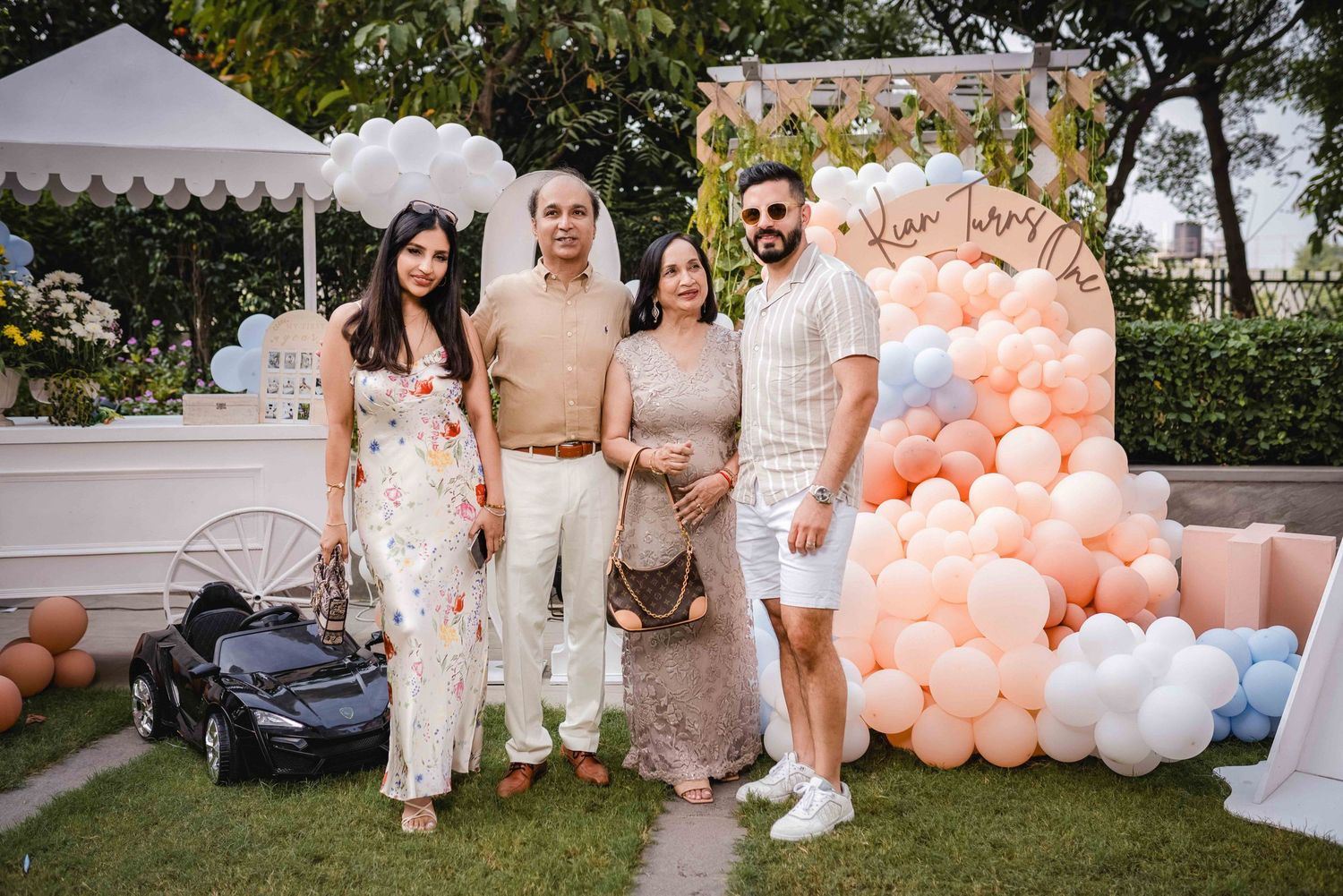 Four people posing together at a festive outdoor party with colorful balloon decorations and a toy car on green grass.