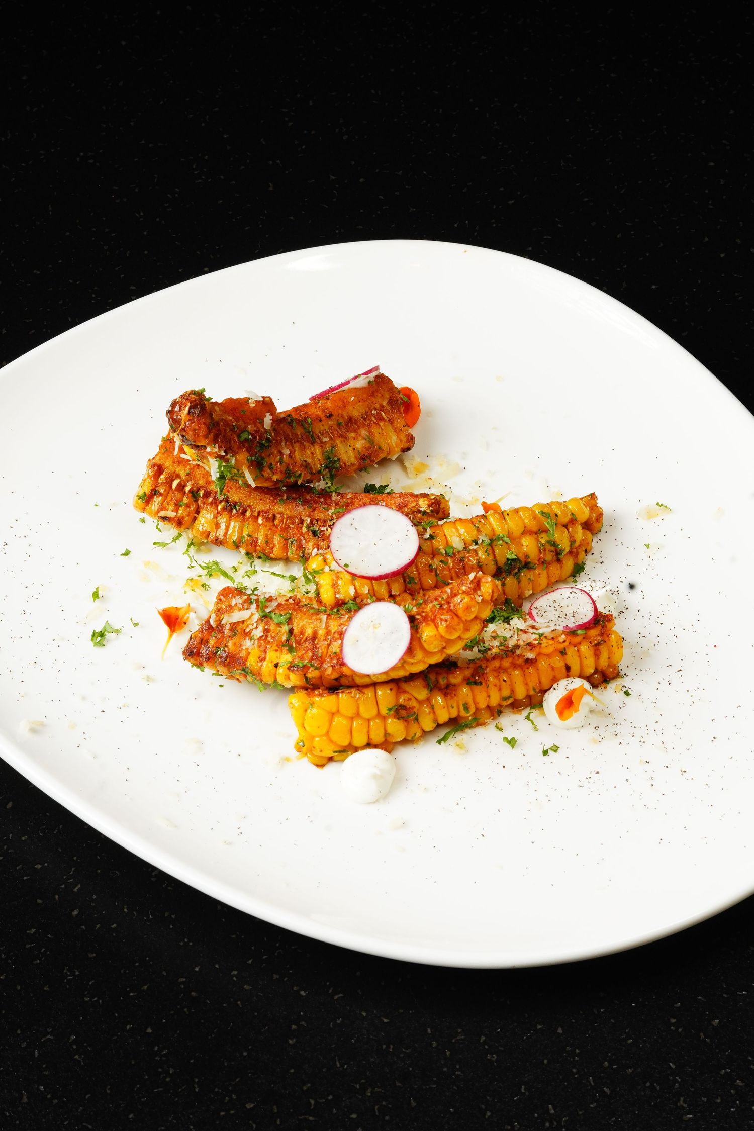 Grilled corn on the cob with spicy seasoning and white sauce drizzle served on white plates in sequence.