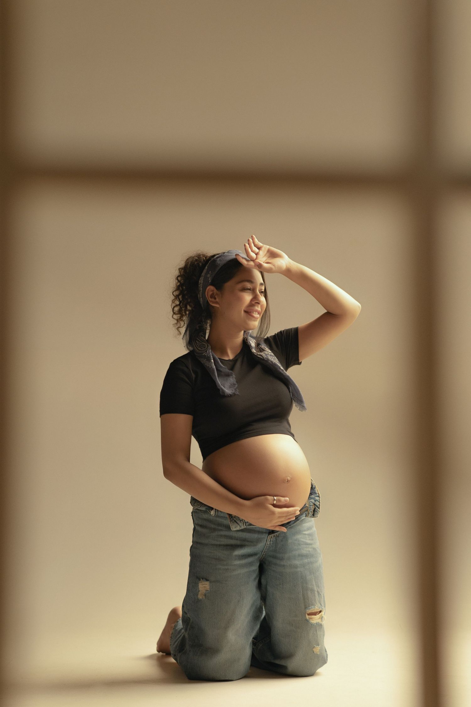 A pregnant woman in a black crop top and ripped jeans kneels while posing against a neutral background.
