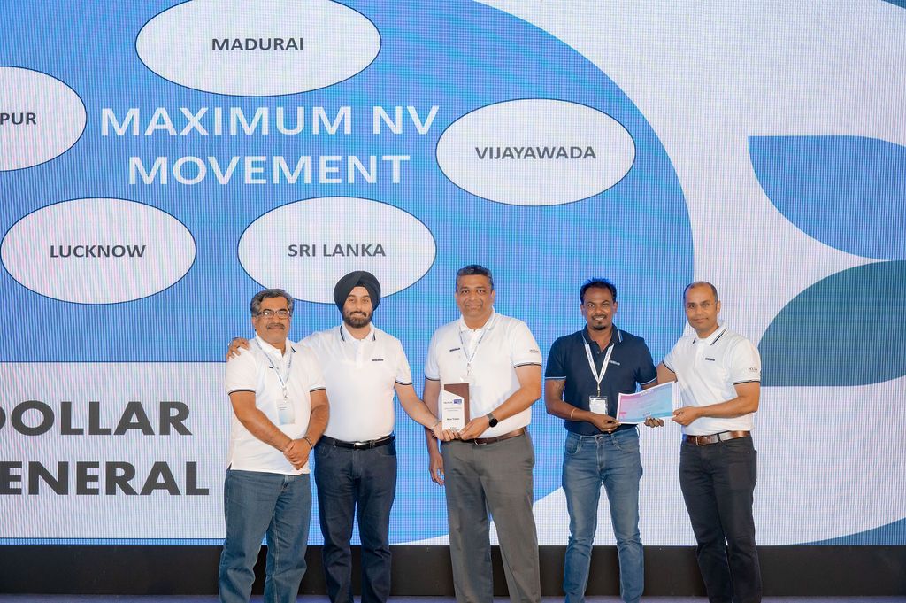 A group of five people in white shirts standing on stage at a corporate event with Maximum NV Movement signage.