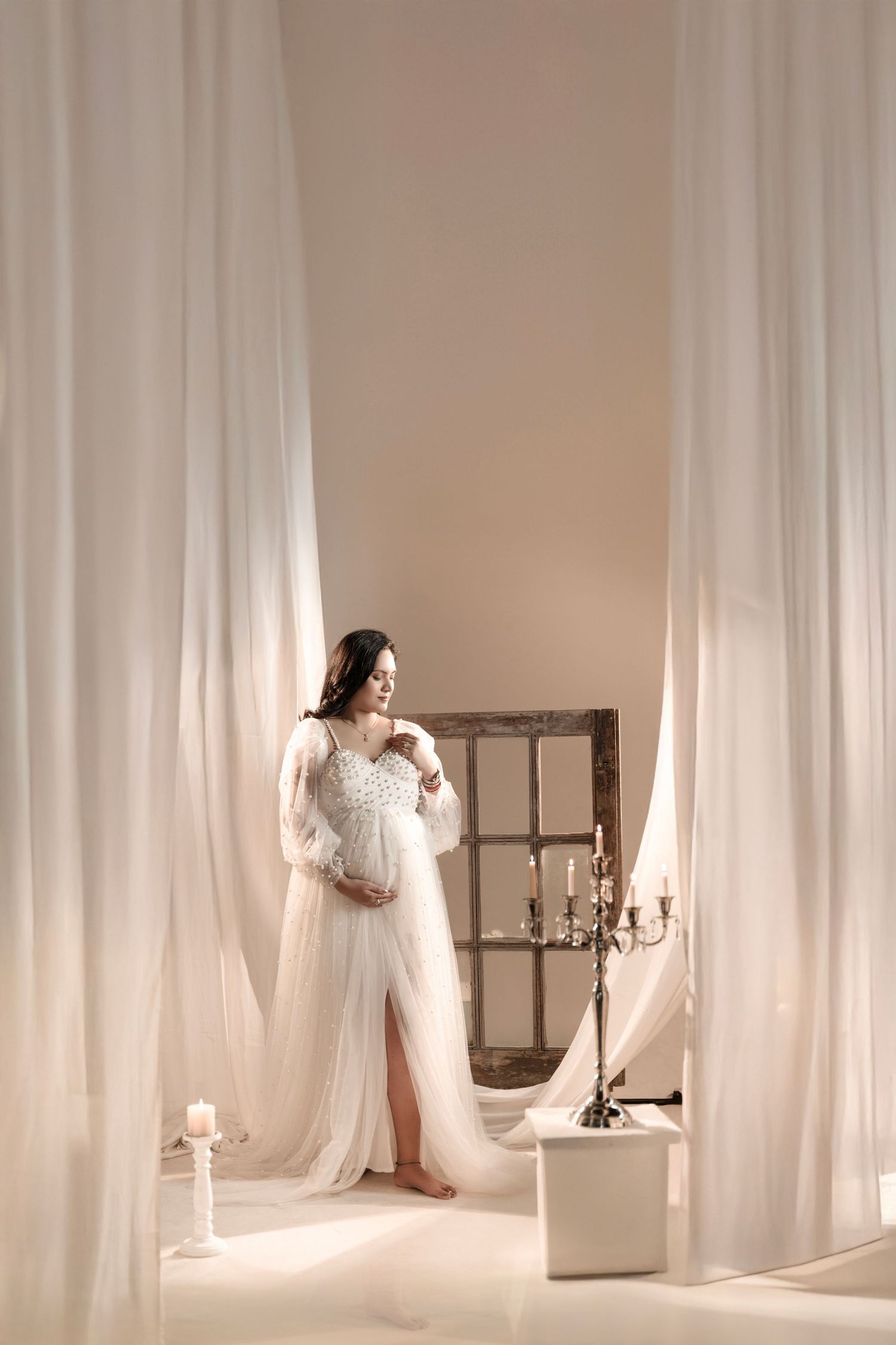 Pregnant woman in elegant white lace gown with flowing tulle skirt poses gracefully in soft, dreamy studio setting.