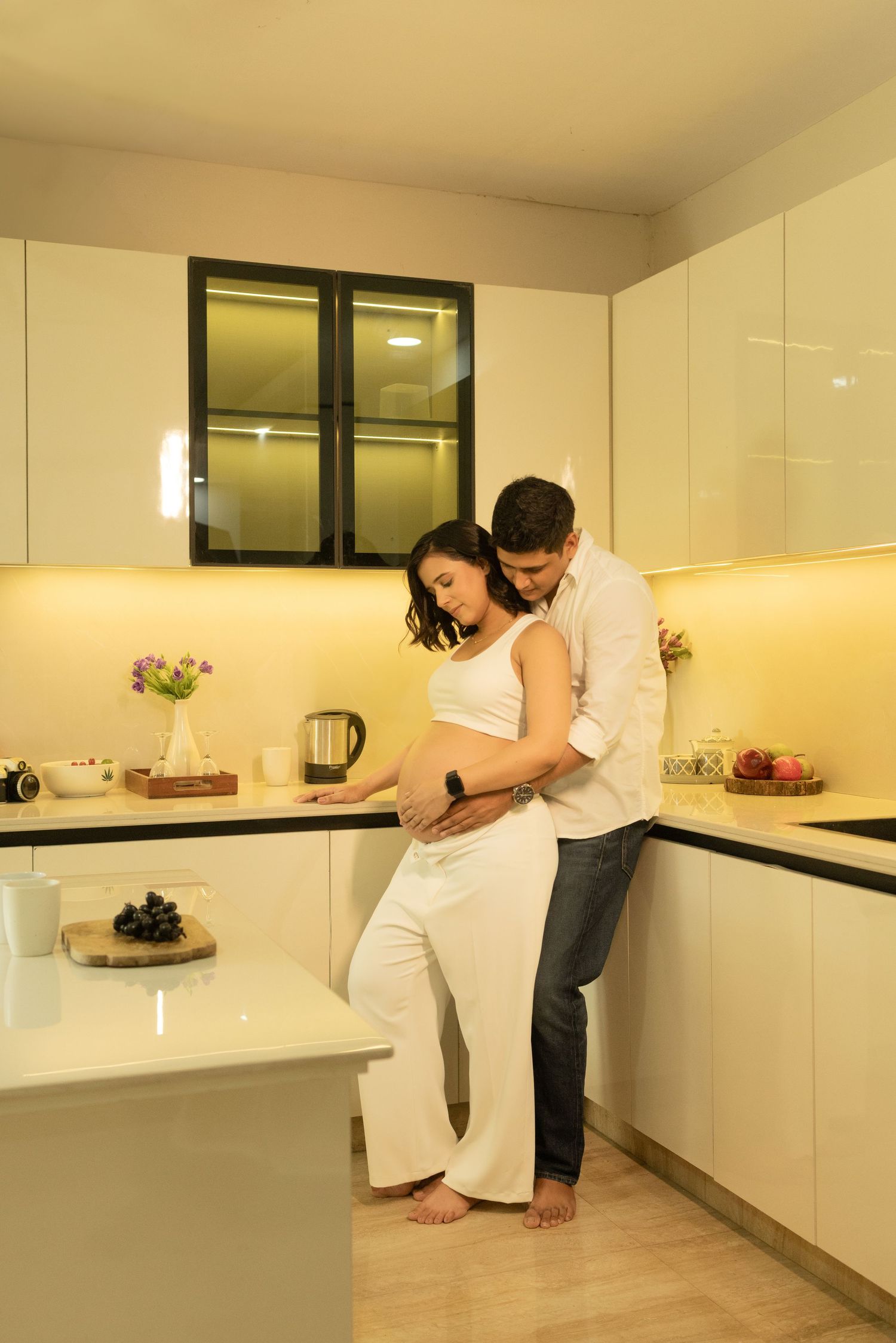 Couple in white kitchen, man embracing pregnant woman from behind tenderly.