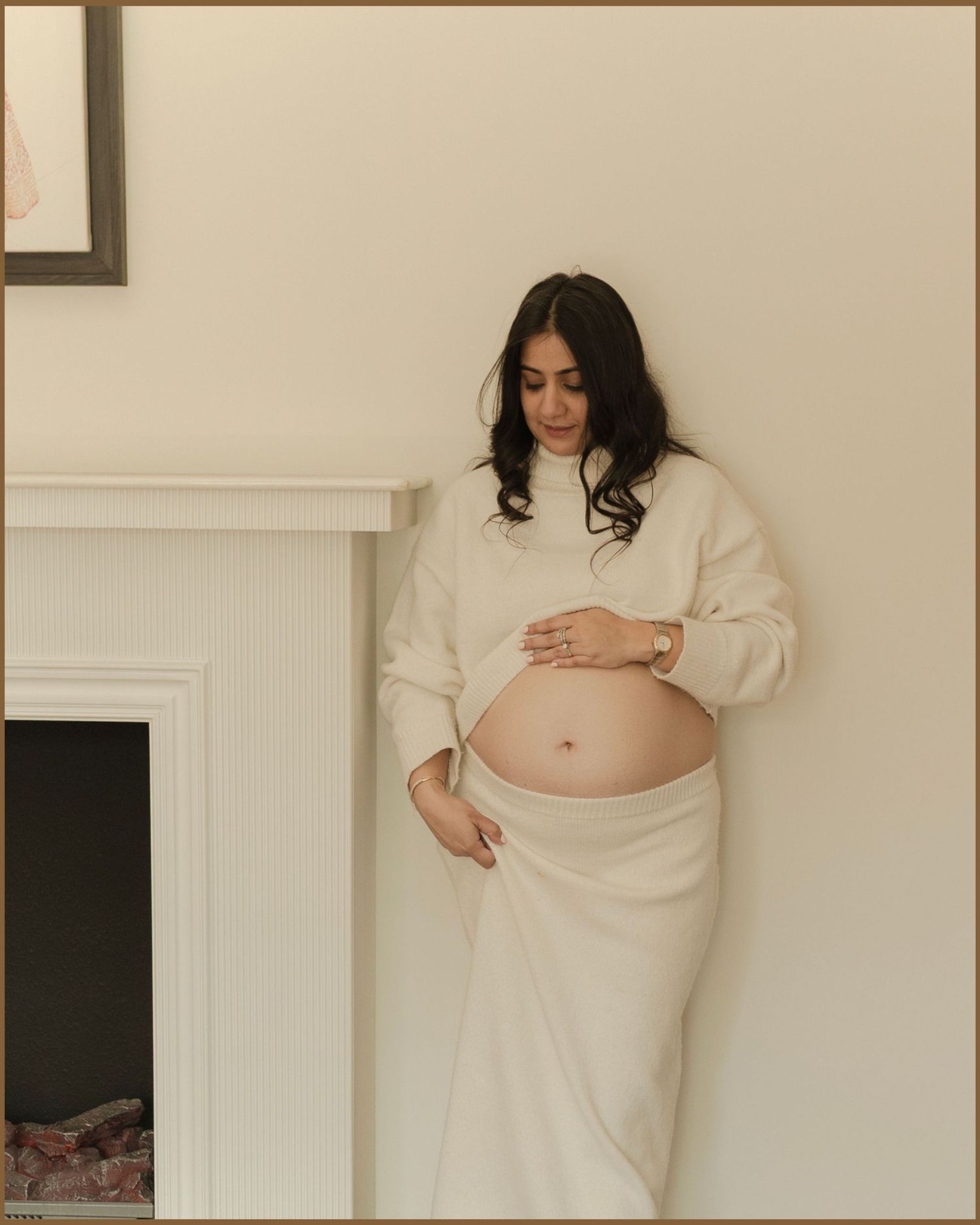 Pregnant woman in cream sweater and draped skirt cradling baby bump, standing by white fireplace in soft neutral tones.