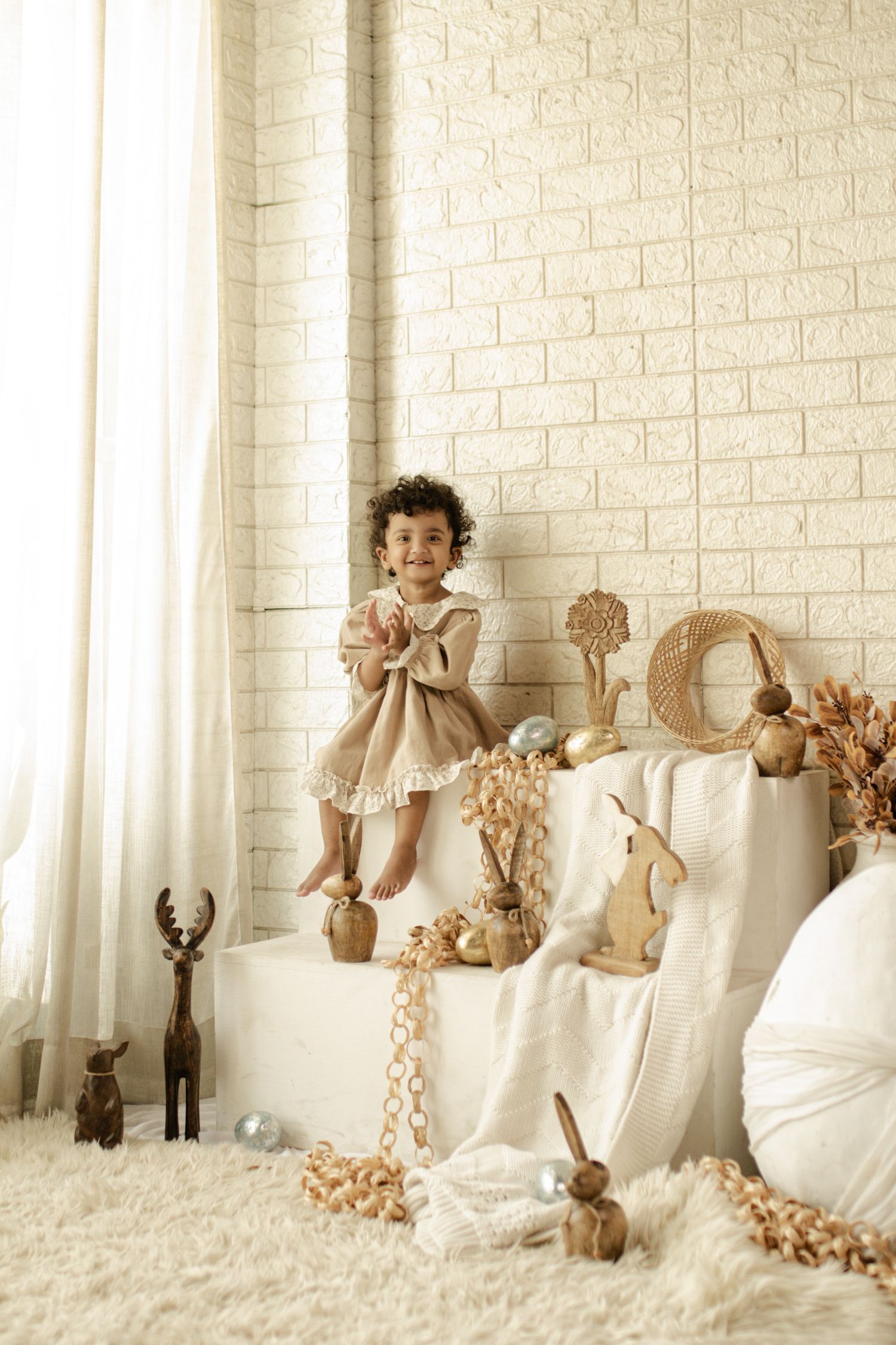 Smiling toddler girl in beige dress standing on white cube surrounded by natural wooden toys and Easter decorations.