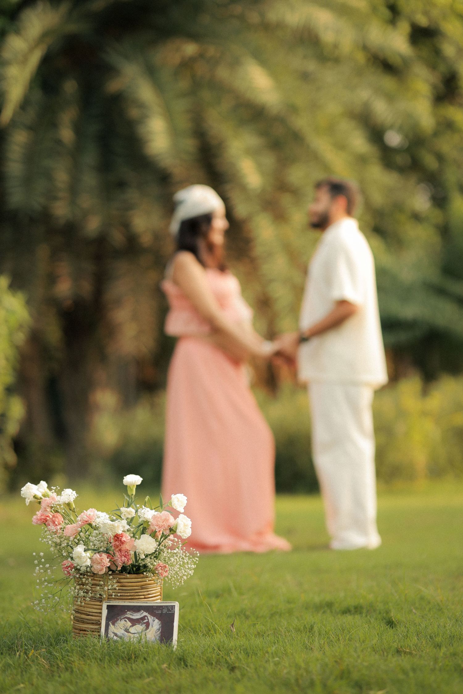 Pregnant couple holding hands in park with floral arrangement and ultrasound photo in foreground.