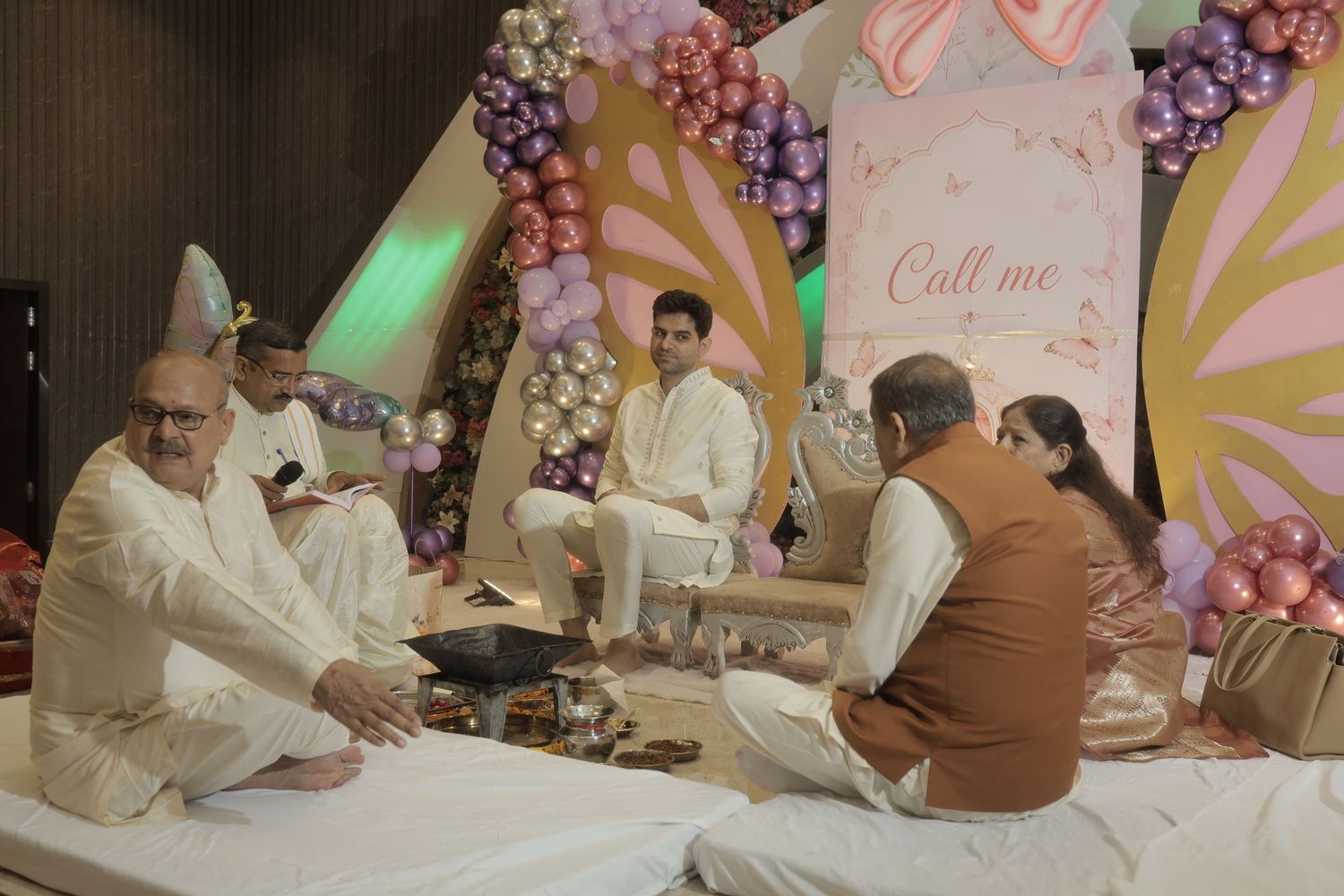 Men in white traditional attire sit on a white-covered floor at a ceremony decorated with colorful balloons and a pastel floral backdrop.