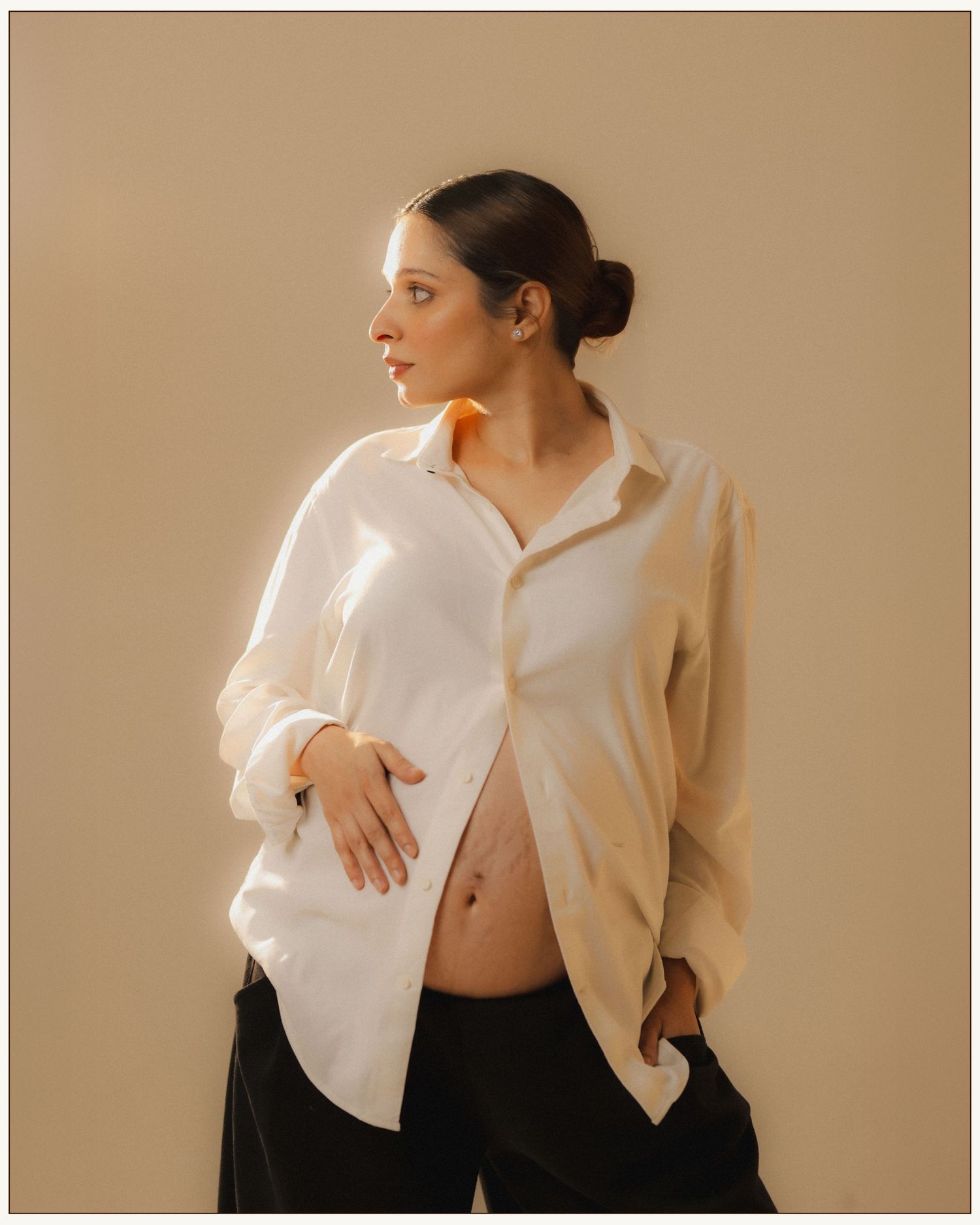 Pregnant woman in white oversized shirt and black pants cradling baby bump, looking sideways against beige background.