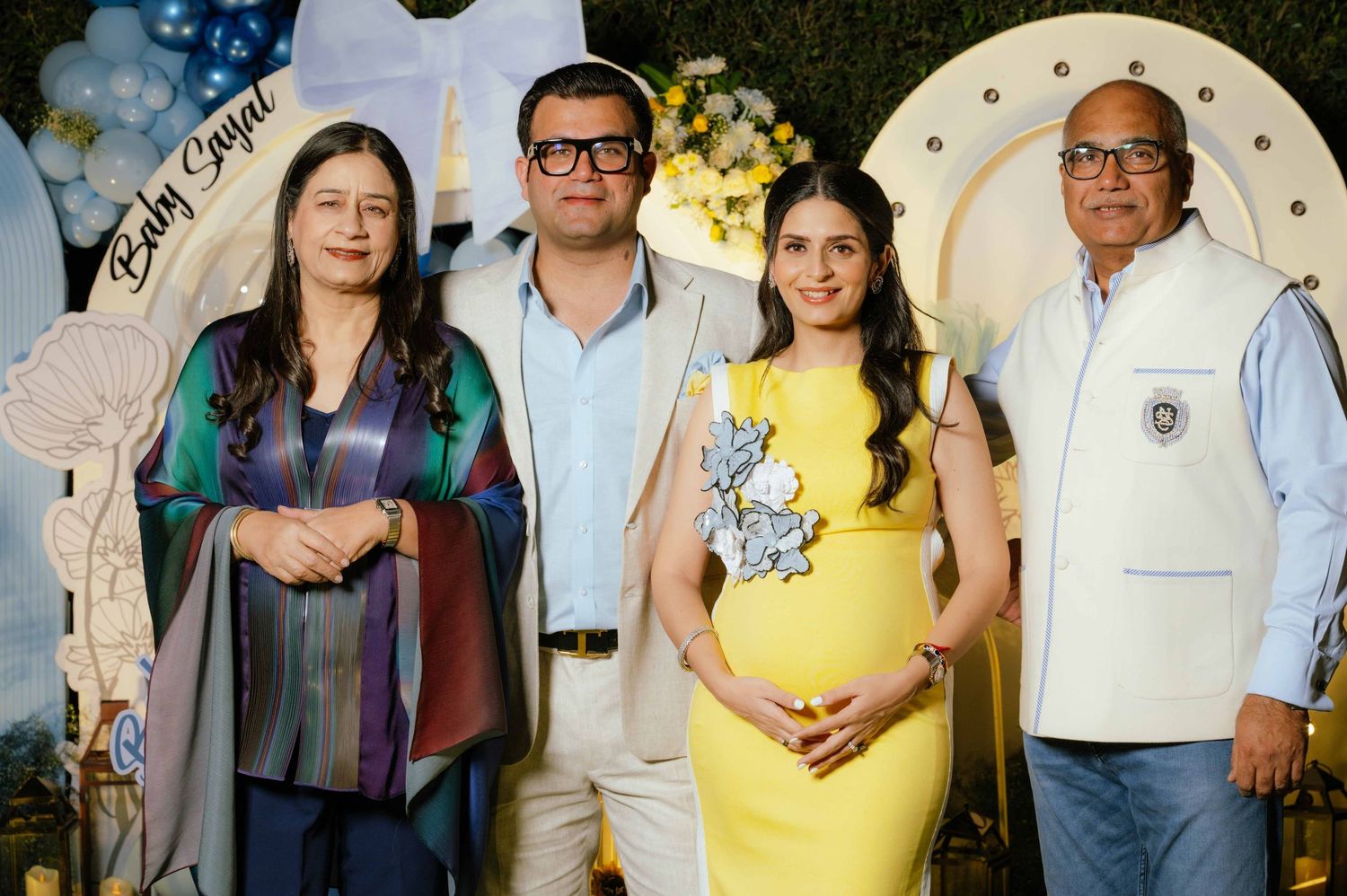 Four people pose together at a baby shower event with blue and white decorations, including a pregnant woman in a yellow dress.