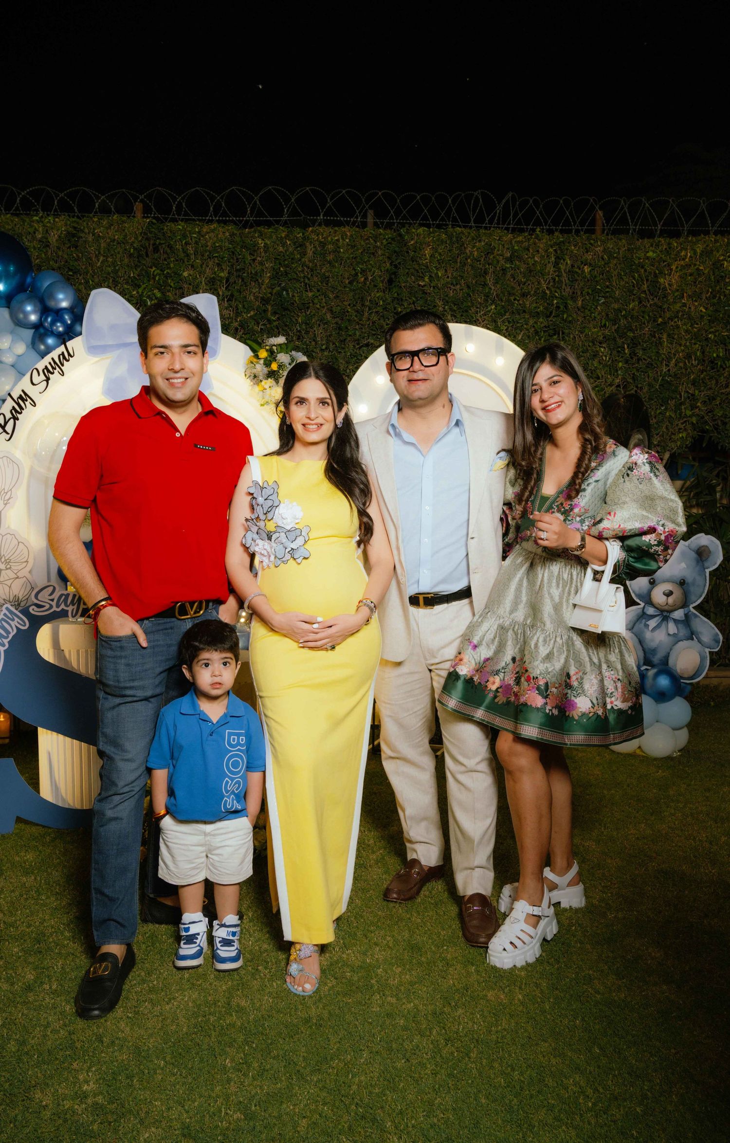 Family posing at a baby shower with a pregnant woman in yellow dress, blue balloon decorations and teddy bear backdrop at night.