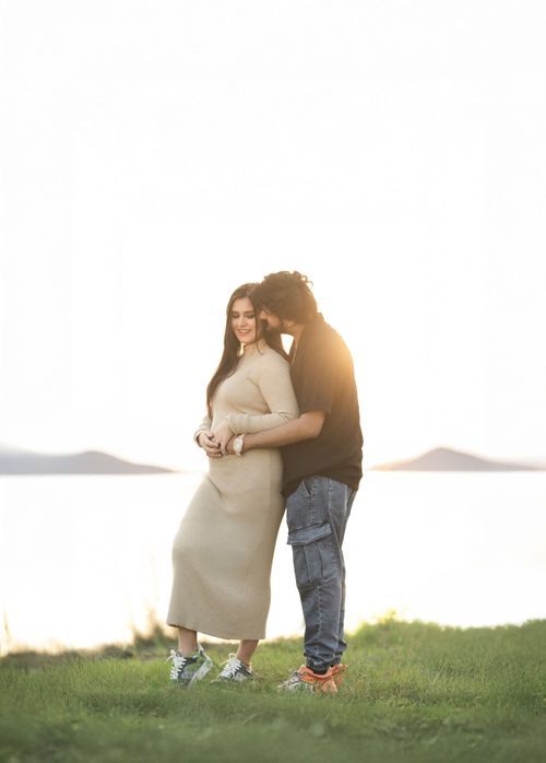 A couple shares an intimate moment on a grassy hillside overlooking a serene lake at sunset.