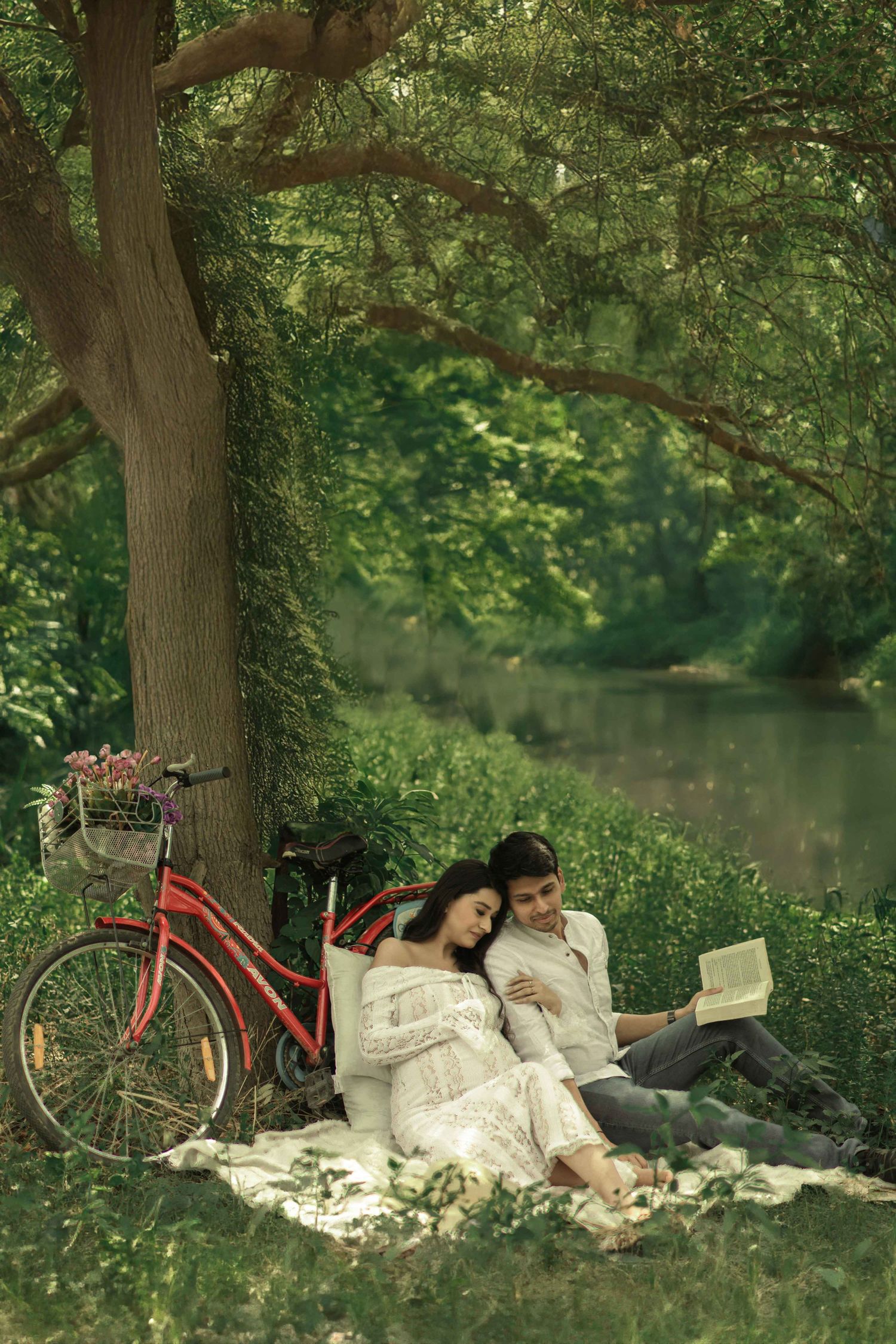 Couple relaxing under a large tree by a river, reading a book on a blanket with a red bicycle nearby.