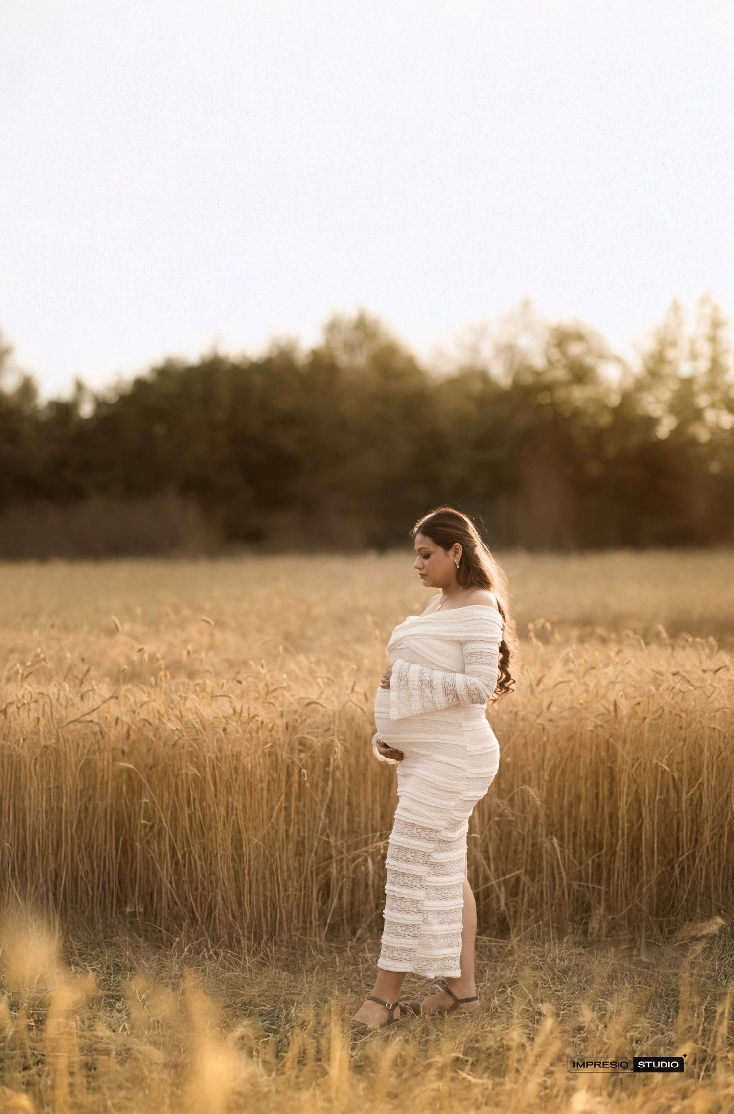 Pregnant woman in white dress standing in golden wheat field during sunset maternity photoshoot.