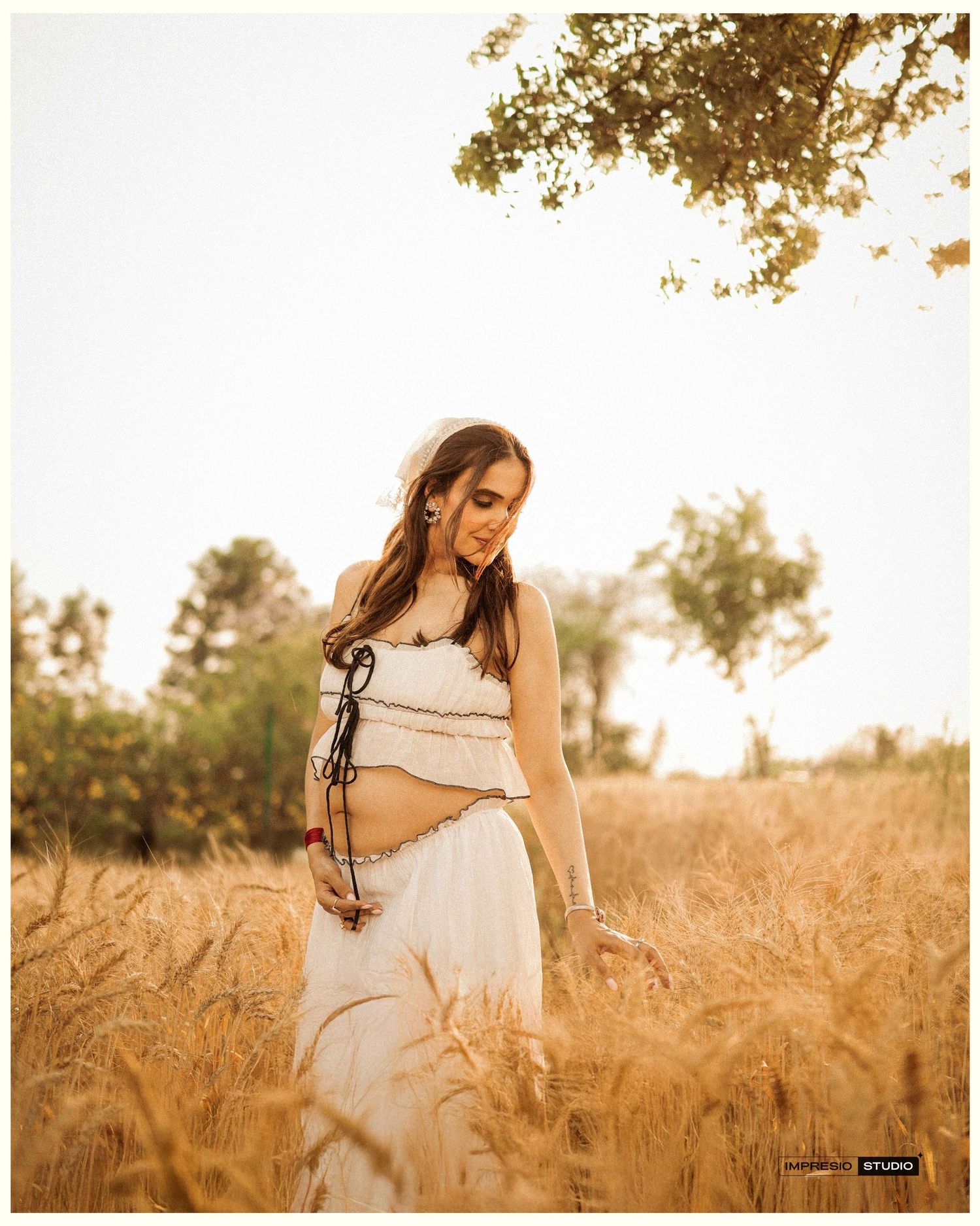 Pregnant woman in white crop top and skirt standing in golden wheat field during sunset.