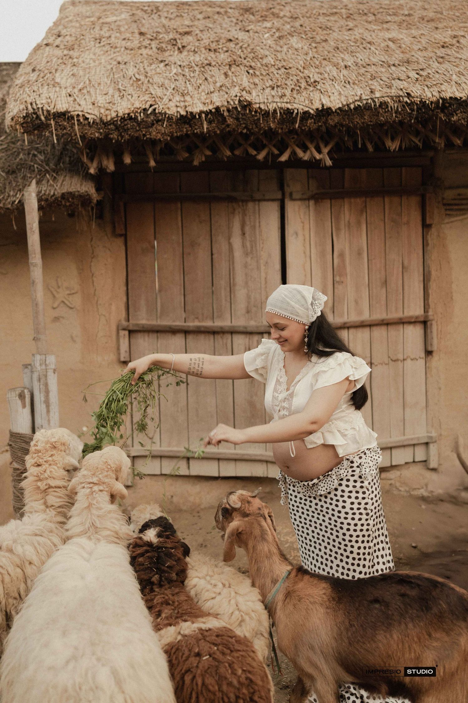Pregnant woman in white top and polka dot skirt feeding green herbs to sheep outside a thatched mud hut.