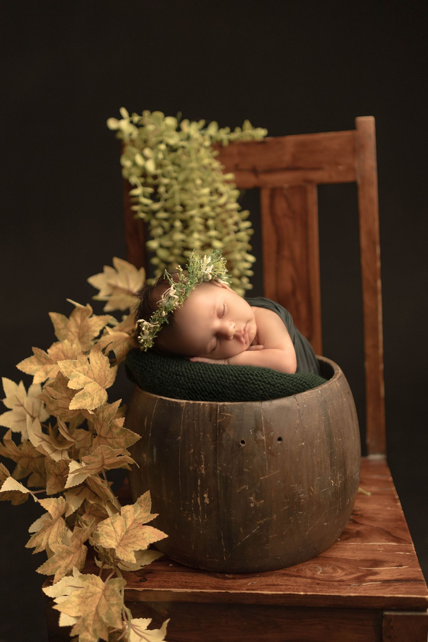 Sleeping newborn baby nestled in a rustic wooden bucket with green wrap and floral crown, surrounded by autumn leaves.