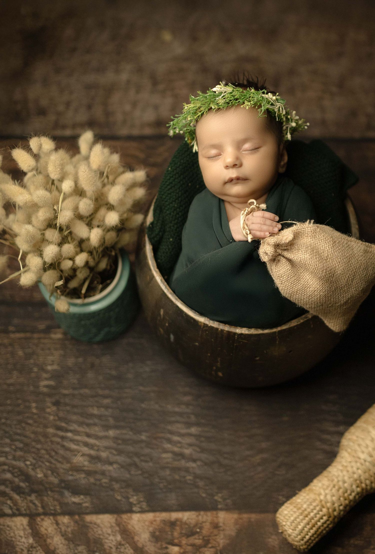 Newborn baby swaddled in green wrap, wearing a floral crown, sleeping in a wooden bowl beside dried flowers.