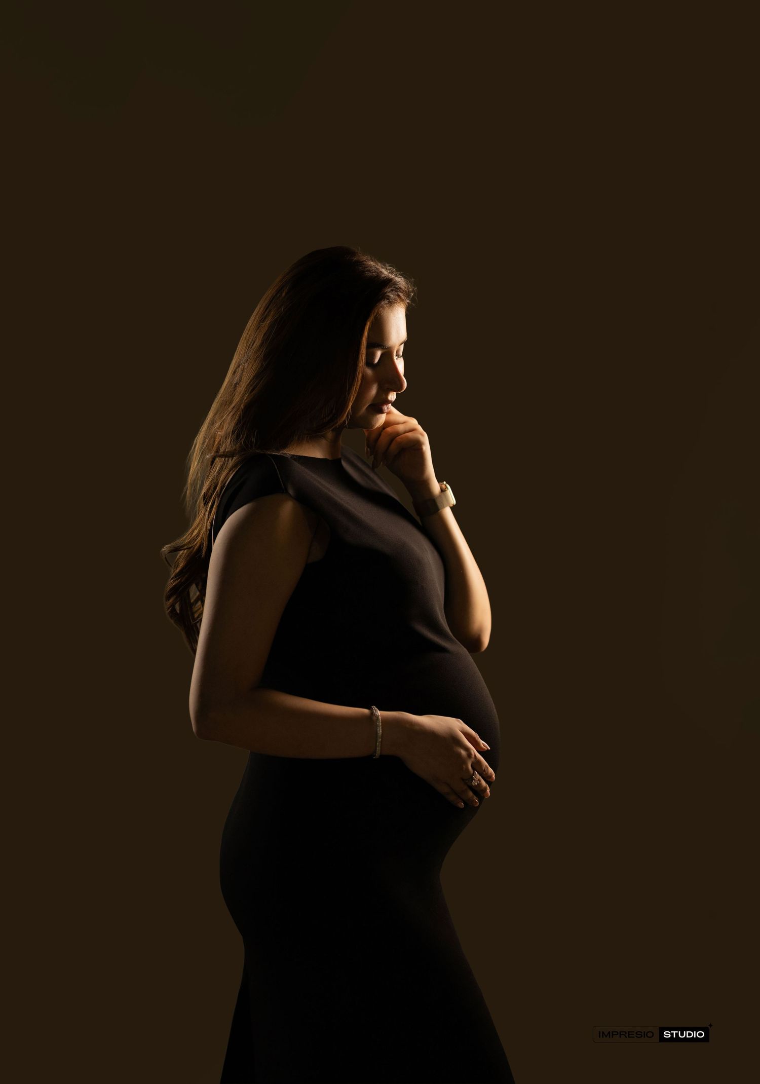 Pregnant woman in black dress poses in dramatic studio lighting, cradling her baby bump in a serene maternity portrait.
