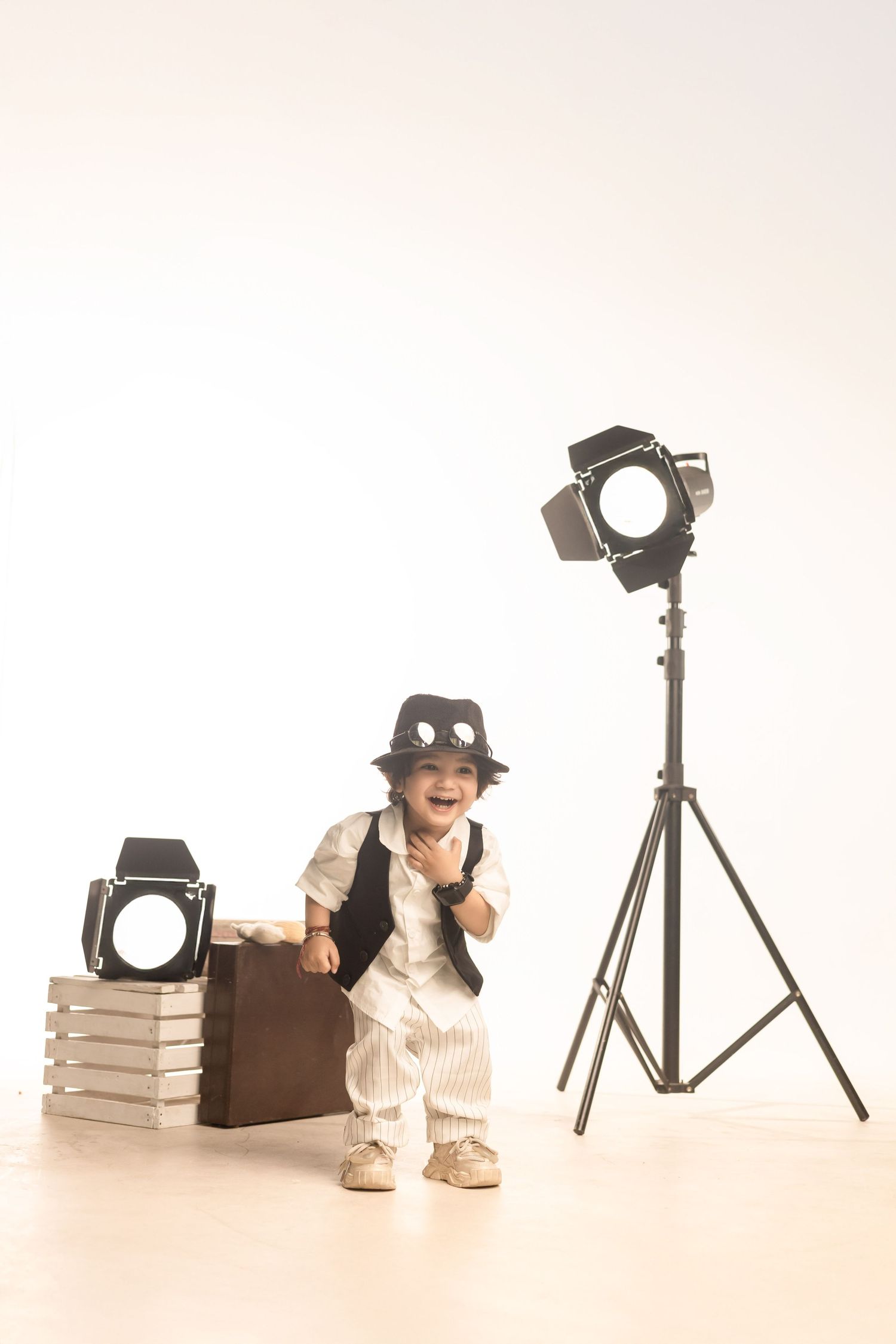 An adorable toddler dressed in a vintage-style suit and hat poses confidently beside a suitcase and studio lights.