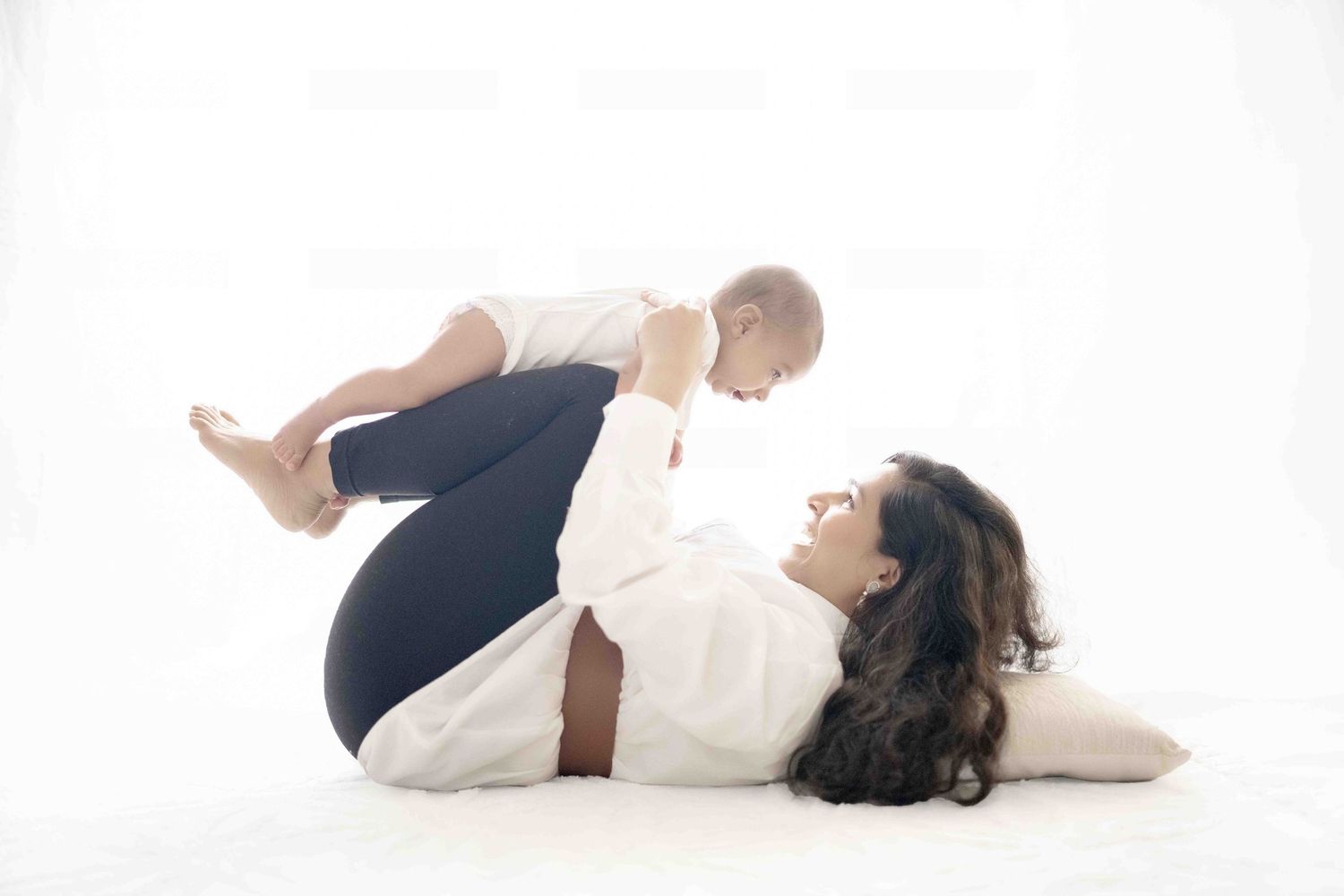 Mother lying on white floor lifting smiling baby in the air during a joyful bonding moment.