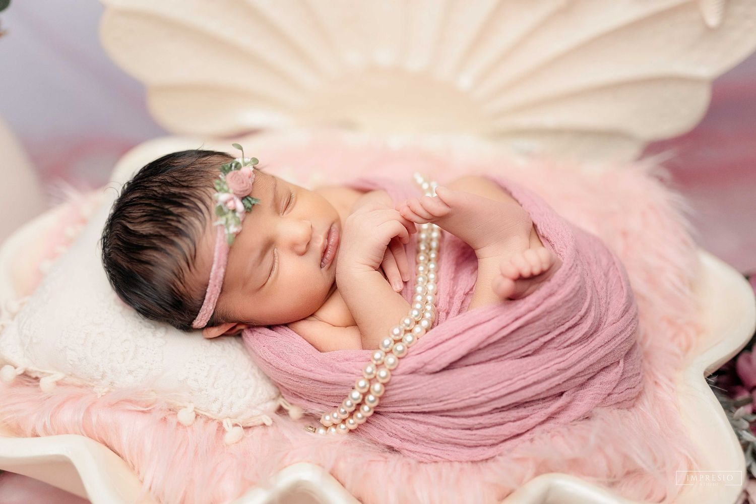 Sleeping newborn baby wrapped in pink swaddle with pearl necklace and floral headband in decorative shell prop.