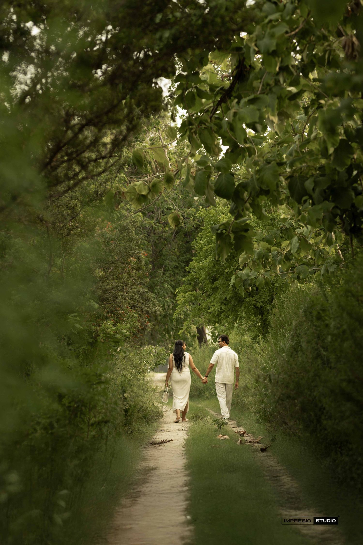 Couple holding hands walking down a lush green tree-lined path in matching white outfits.