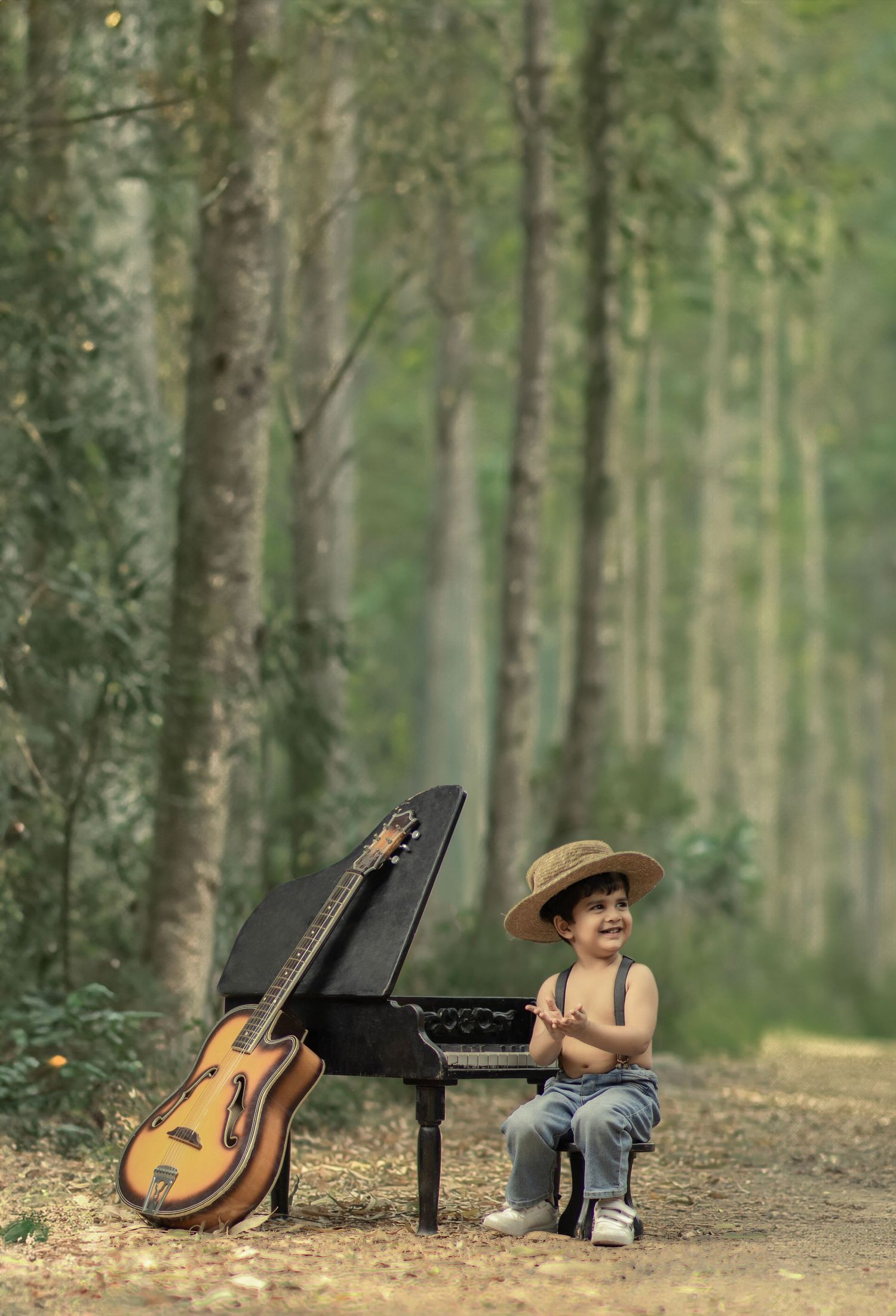 A cheerful toddler in a straw hat sits by a mini grand piano with a guitar leaning against it in a forest path.