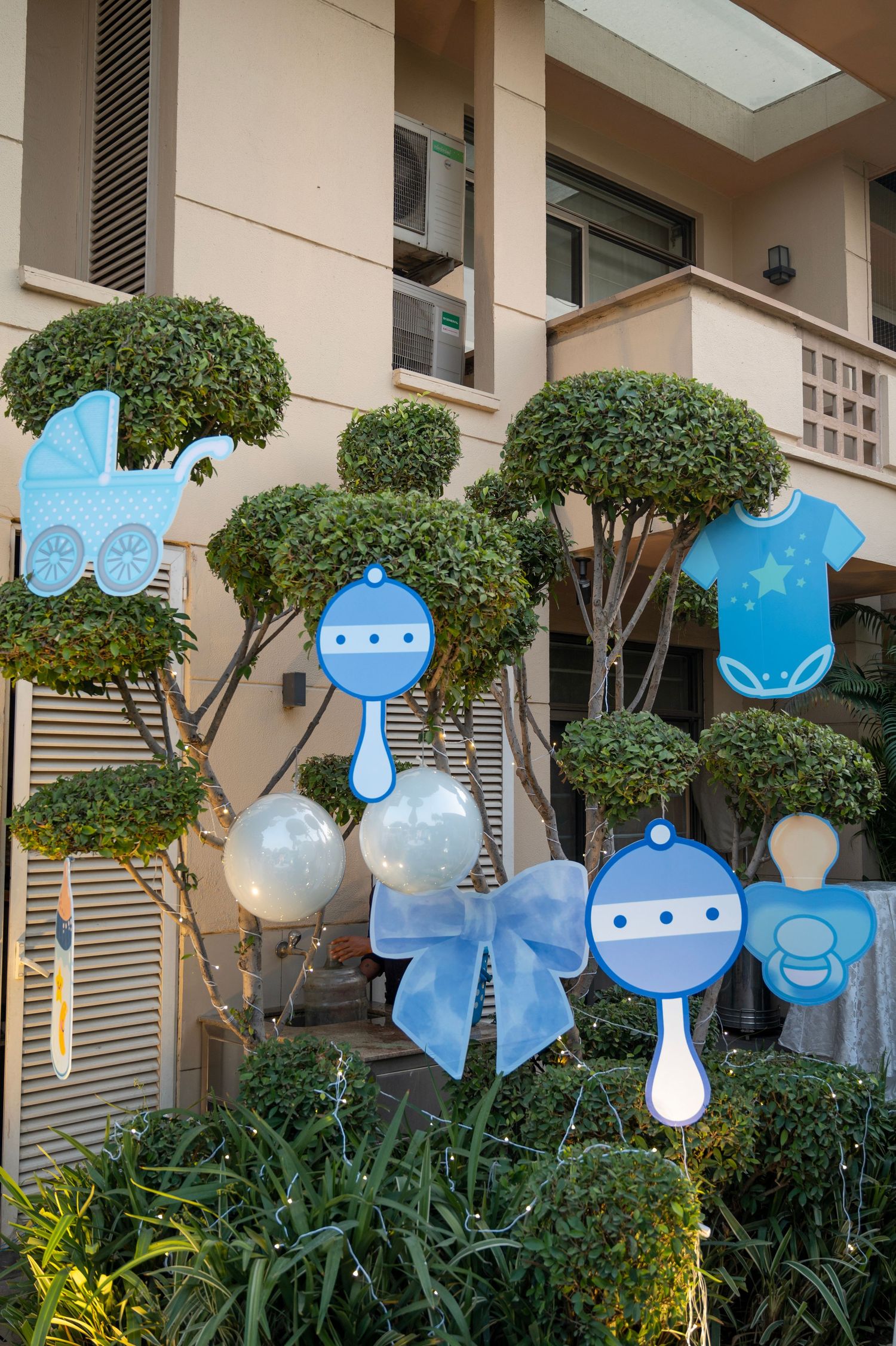 Blue baby shower yard decorations featuring rattles, onesie, pacifier, stroller cutouts, and balloons in front of a building.