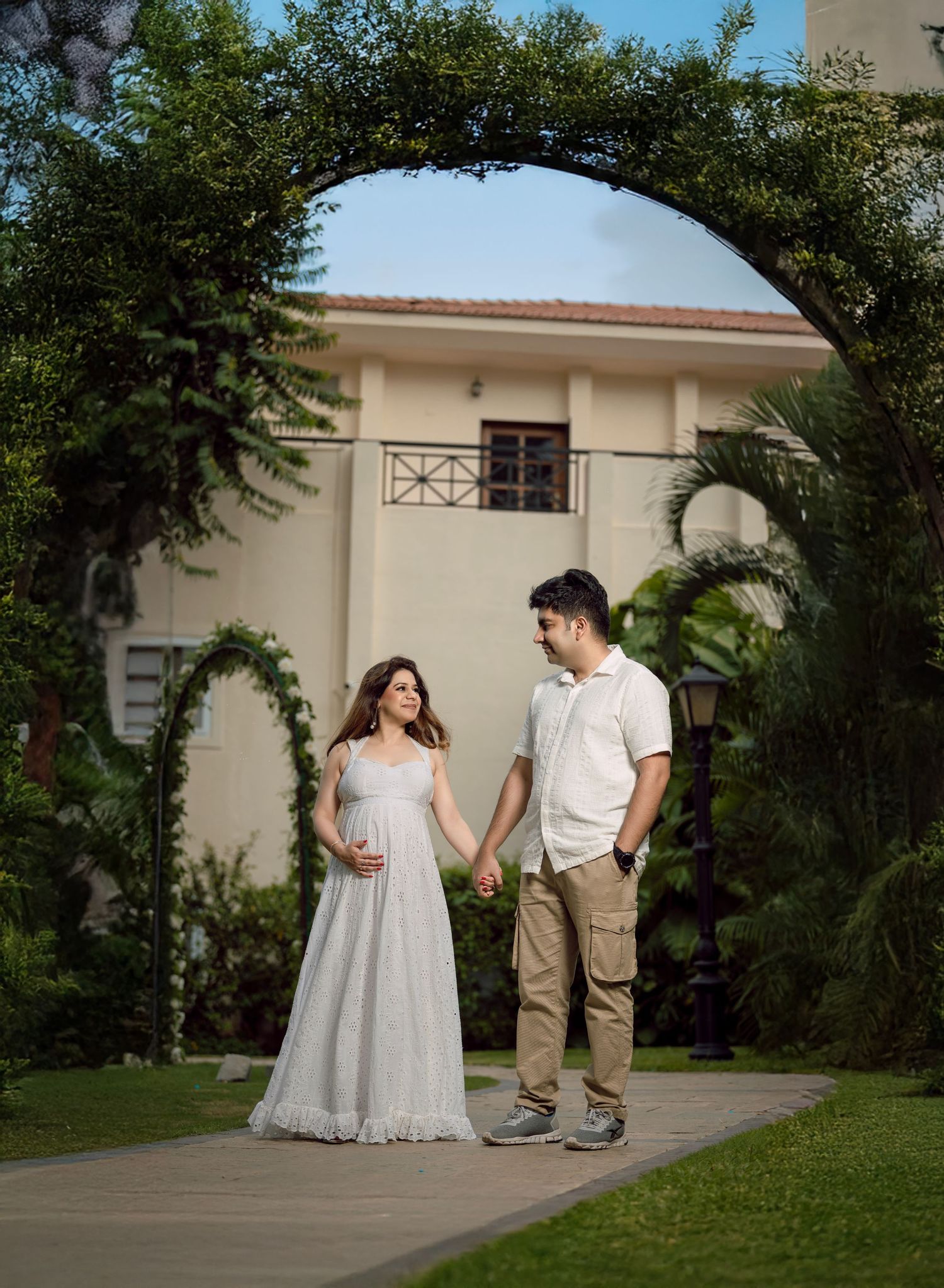 Pregnant couple holding hands under a green arch in a garden, woman in white gown, man in beige outfit.