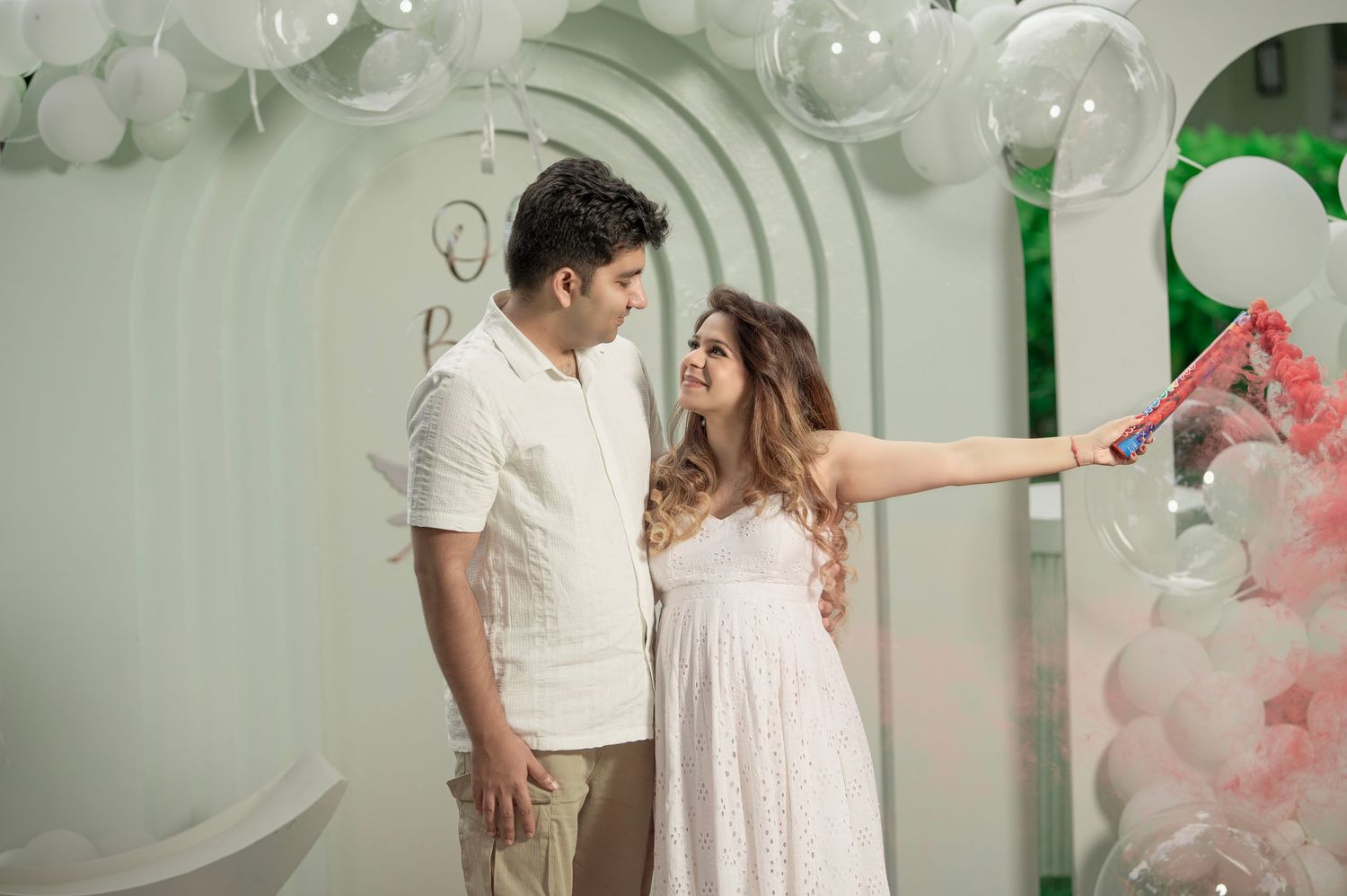 Pregnant woman in white dress with partner at gender reveal party with pink smoke and balloons.