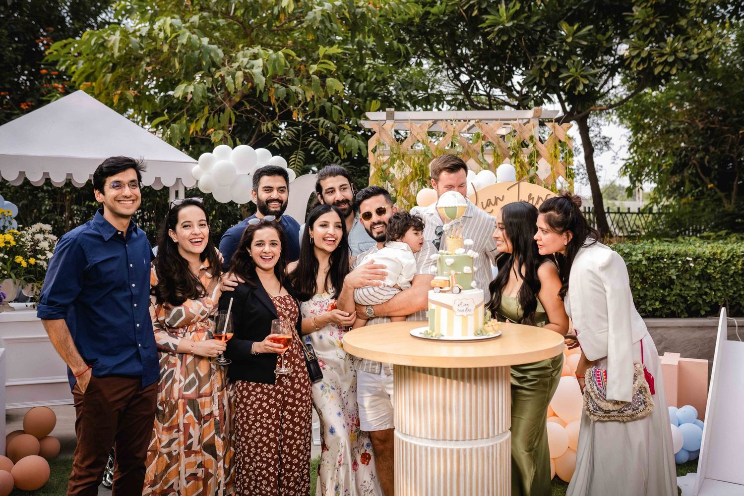 Group of friends celebrating at an outdoor party with a decorated cake table and festive backdrop.