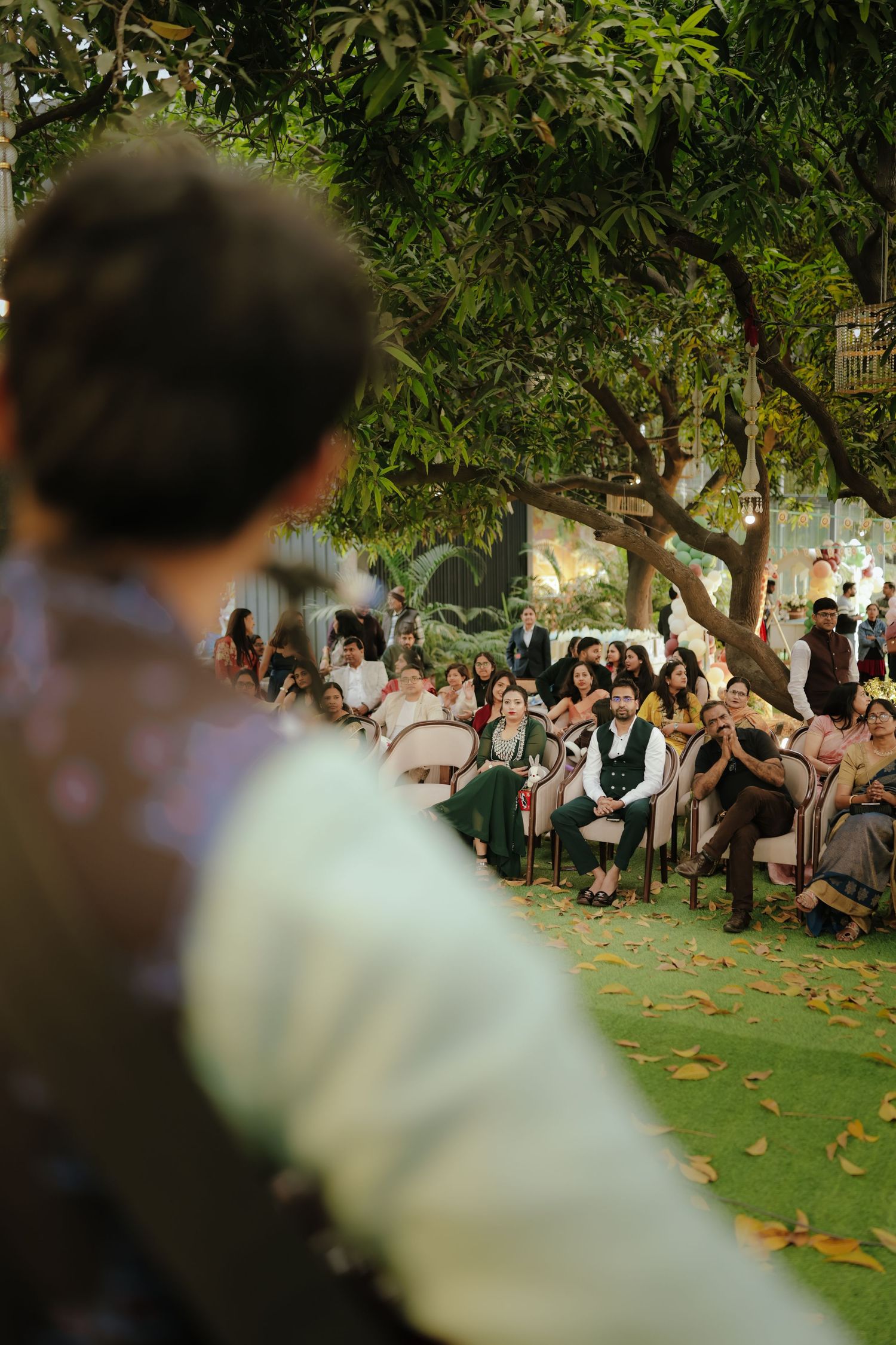Outdoor wedding ceremony with guests seated under lush green trees, viewed from behind the groom.