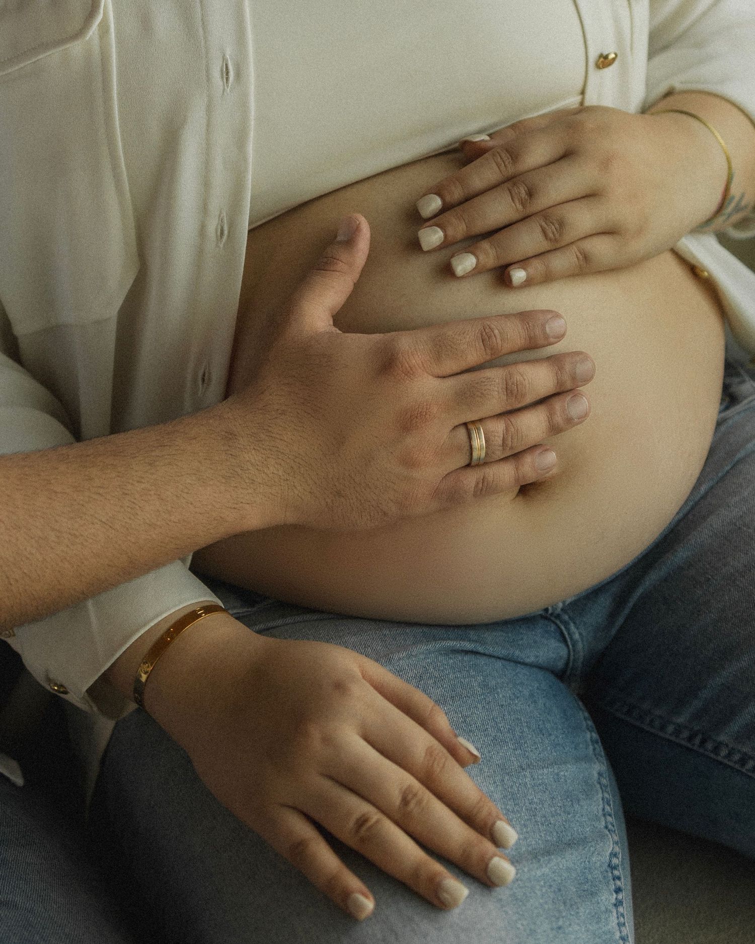 Couple tenderly cradling pregnant belly, woman wearing white shirt and jeans, both adorned with gold jewelry.
